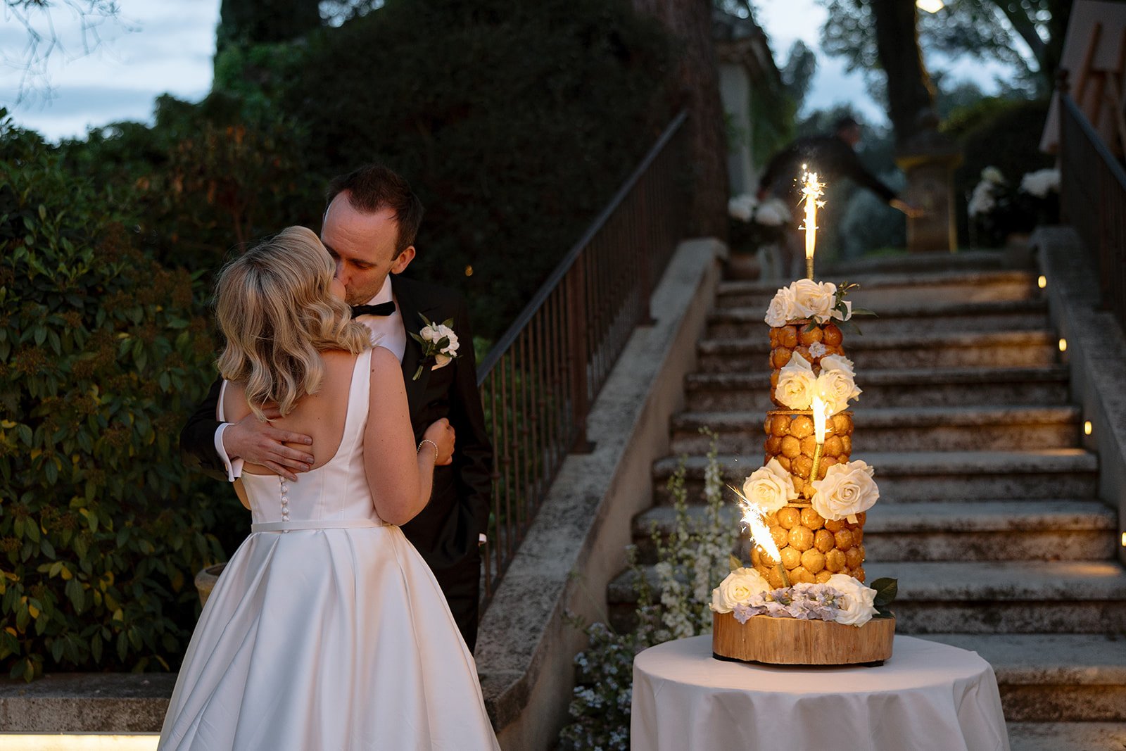 A bride and groom embrace in front of outdoor stairs beside a tall, decorated croquembouche wedding cake with a lit sparkler on top.