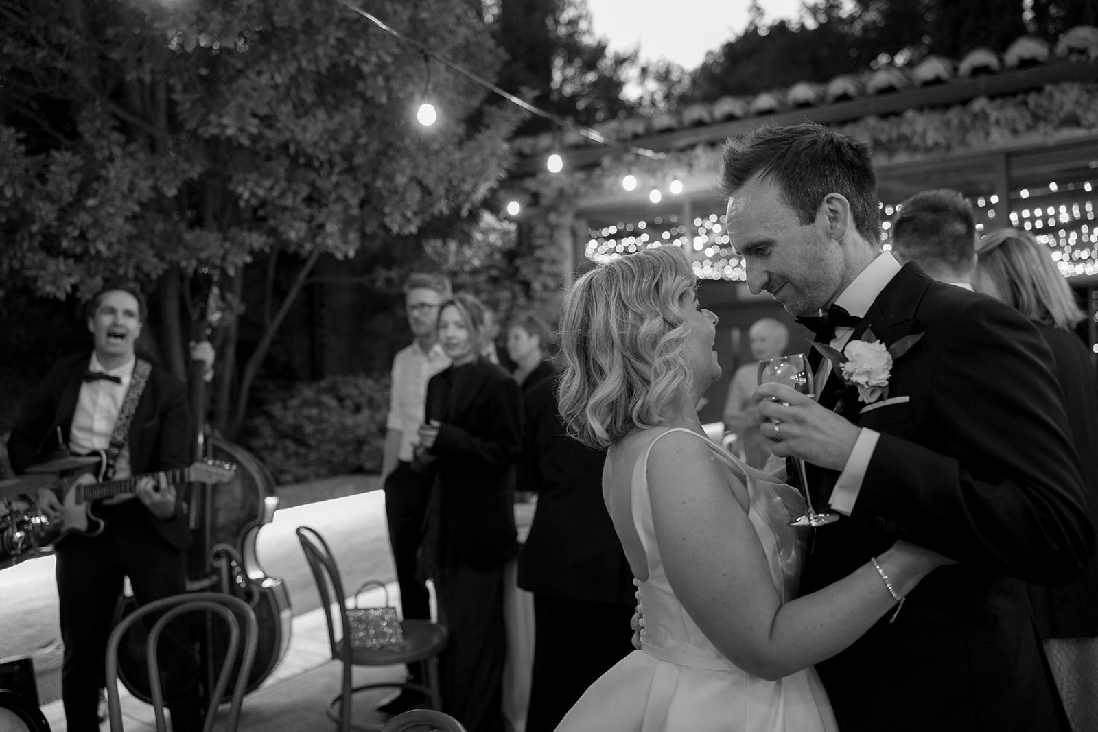 A bride and groom dance closely at their wedding reception while guests watch and a musician plays guitar in the background.