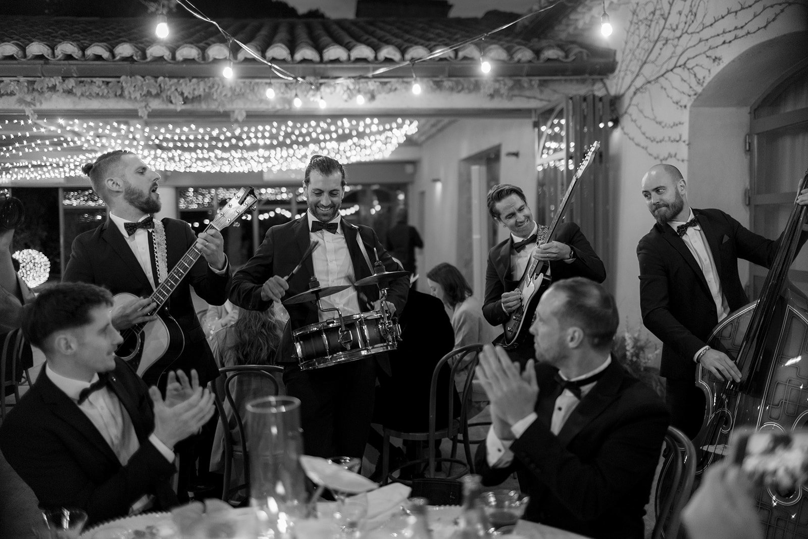 A black and white photo of a band in suits performing with guitar, snare drum, electric guitar, and double bass at a festive event, while seated guests clap and watch. An elegant french wedding.