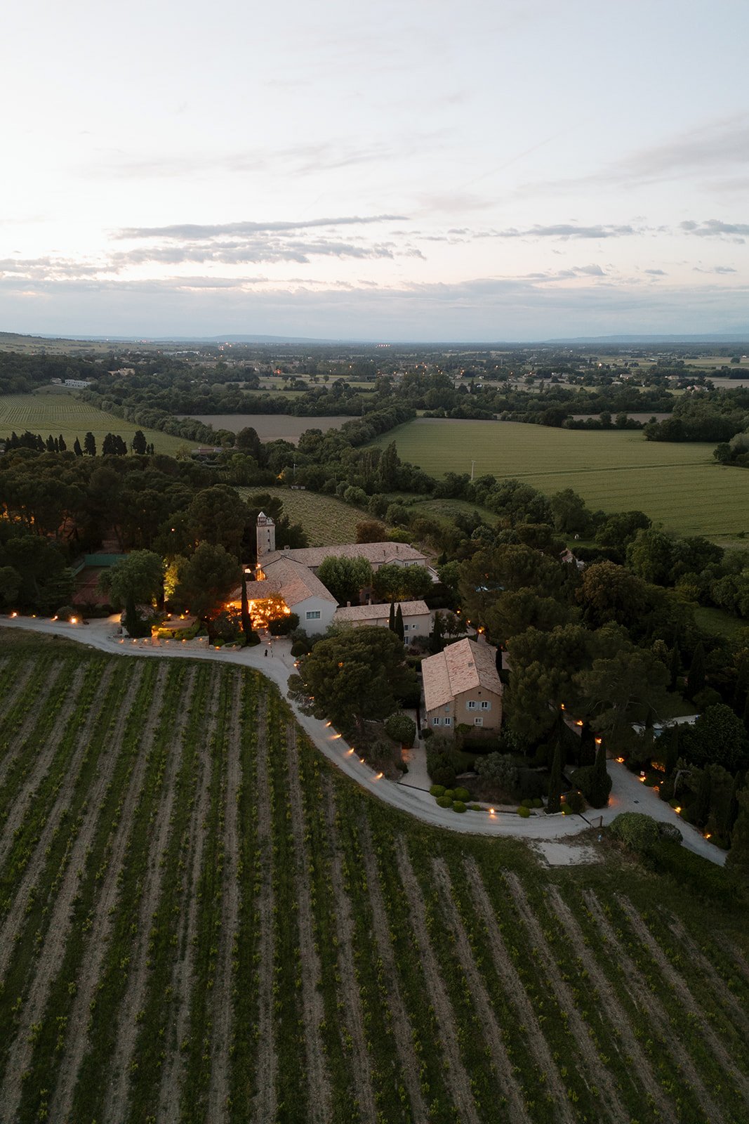 Aerial view of a rural estate with several buildings surrounded by vineyards and farmland at dusk, with landscape stretching into the distance. An elegant french wedding.