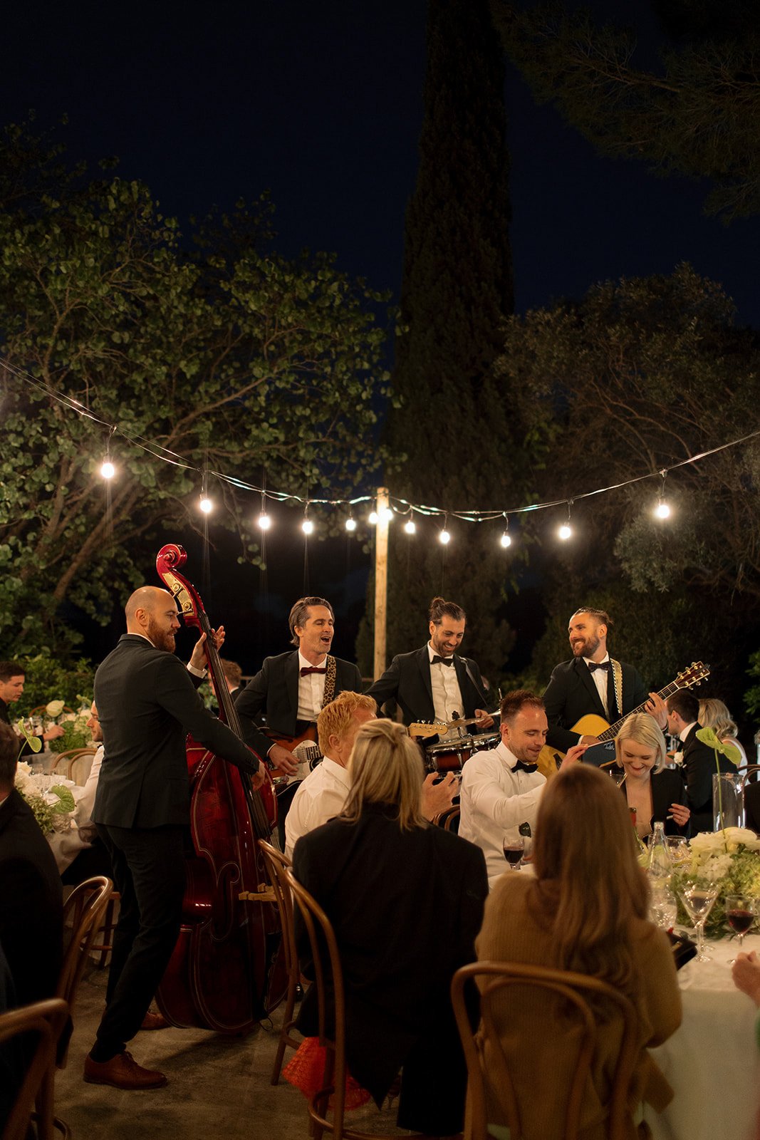 A string quartet in tuxedos performs outdoors at night for seated dinner guests under string lights, surrounded by trees.