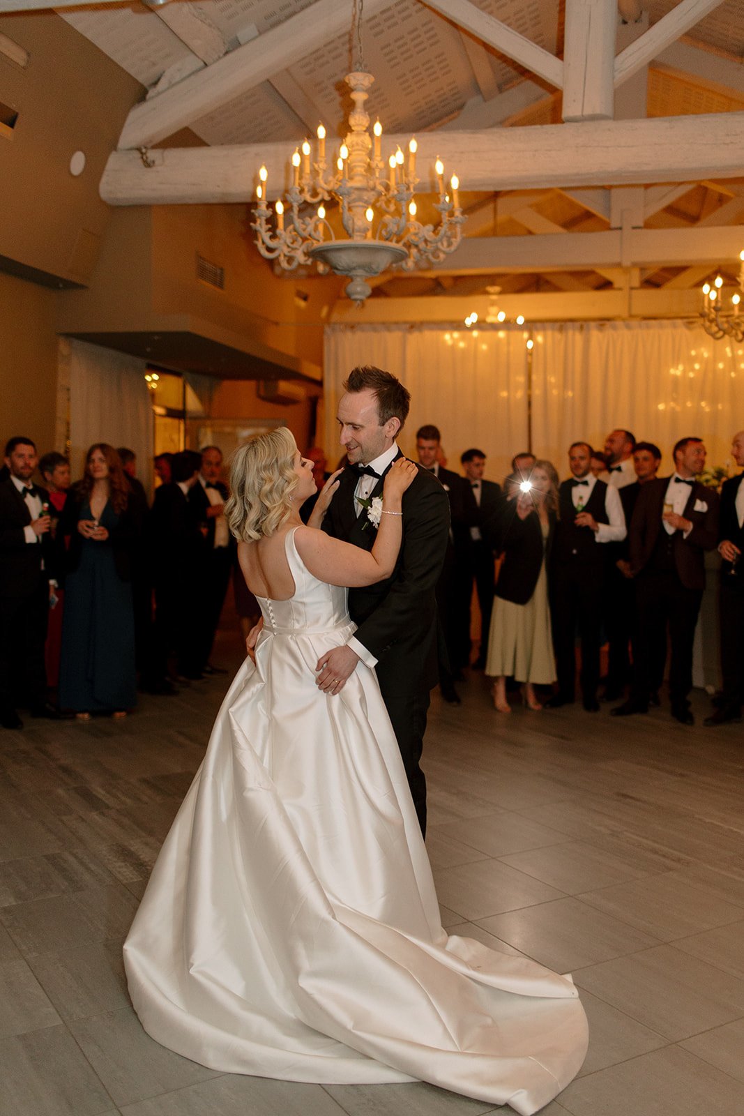A bride and groom share their first dance in a warmly lit banquet hall, surrounded by guests dressed in formal attire.