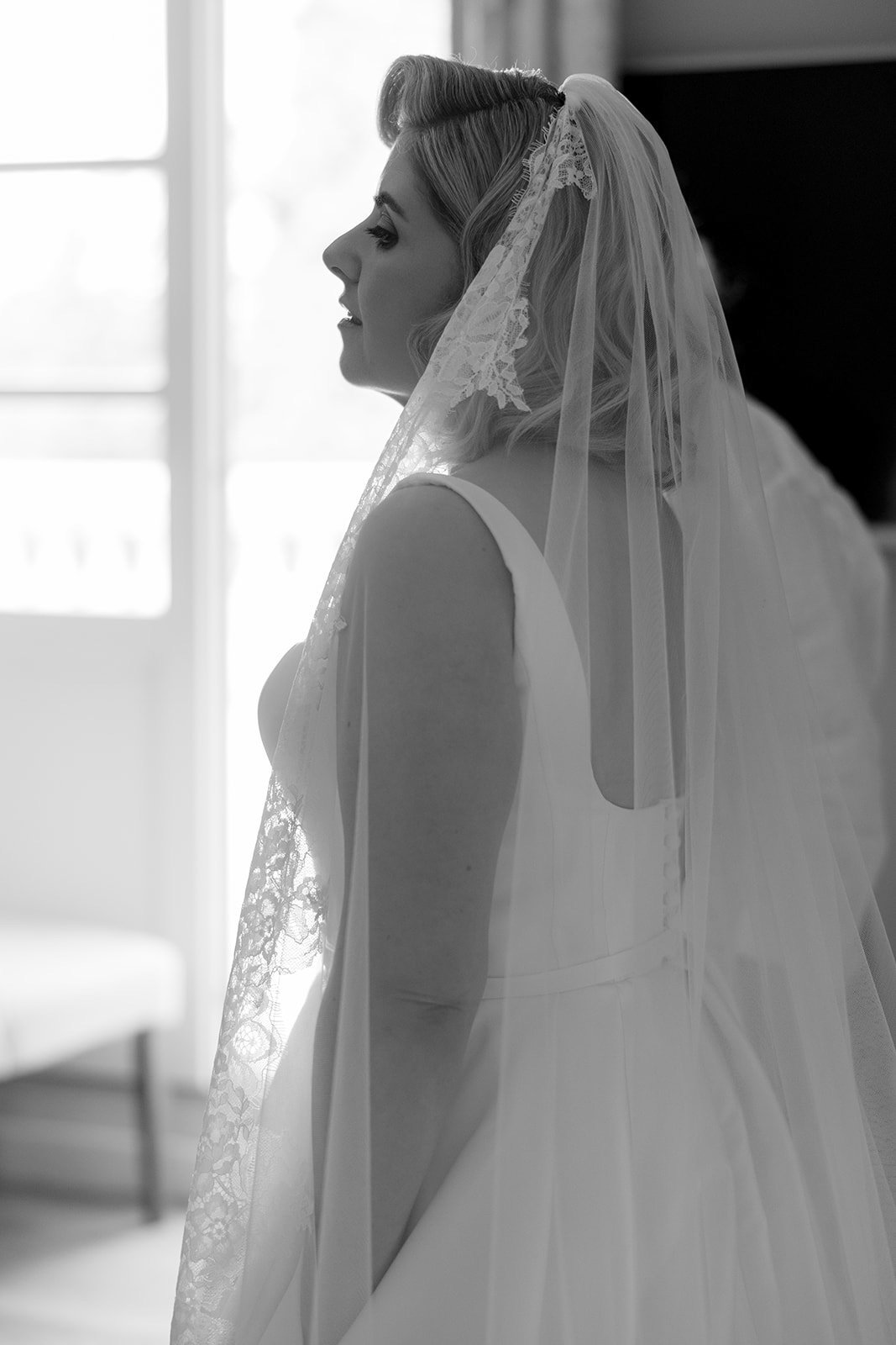 A bride in a white dress and lace veil stands indoors, lit by natural light from a nearby window.