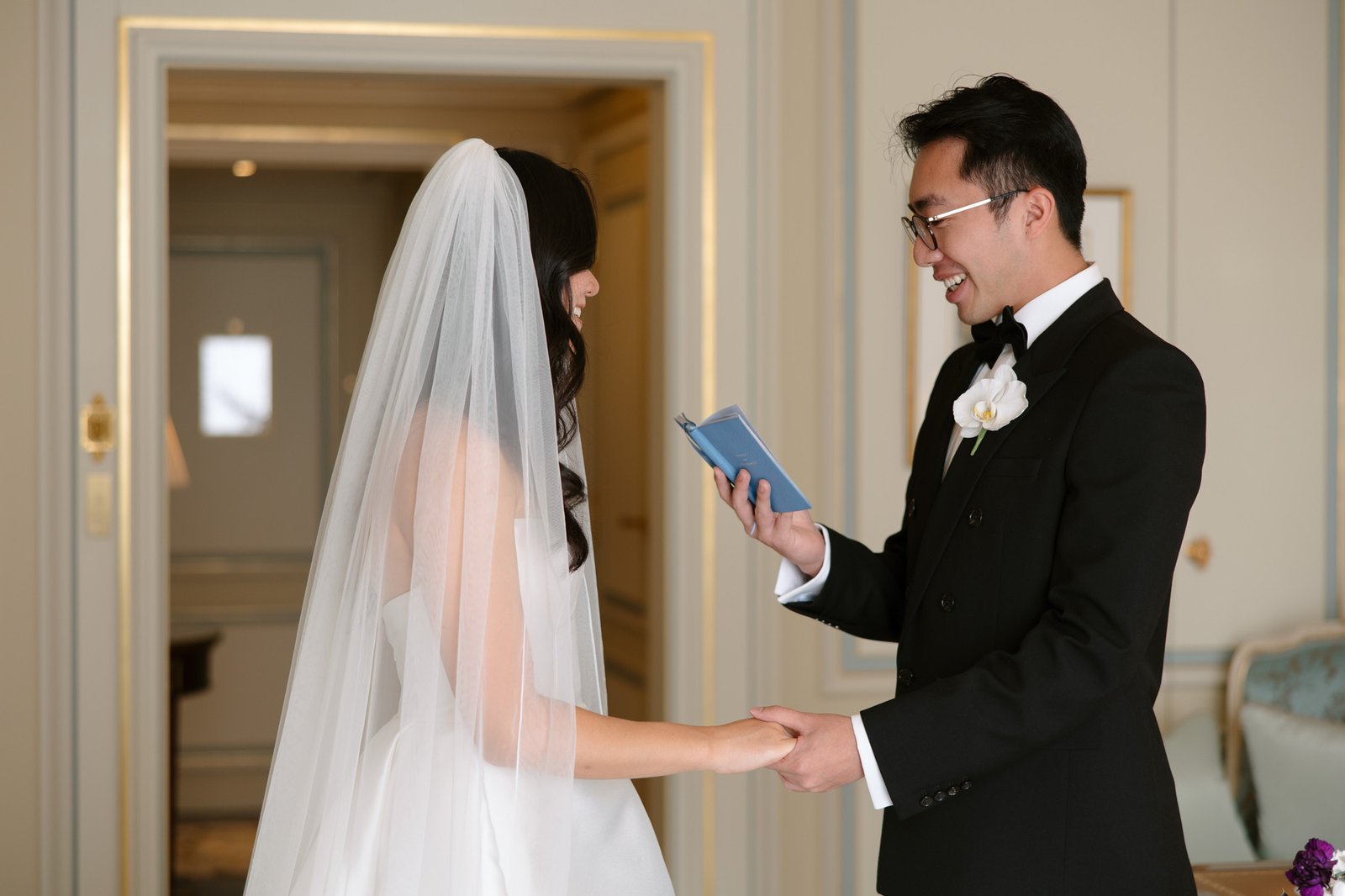 A bride and groom stand indoors, holding hands. The groom reads from a small blue notebook while the bride, wearing a veil and white dress, listens.