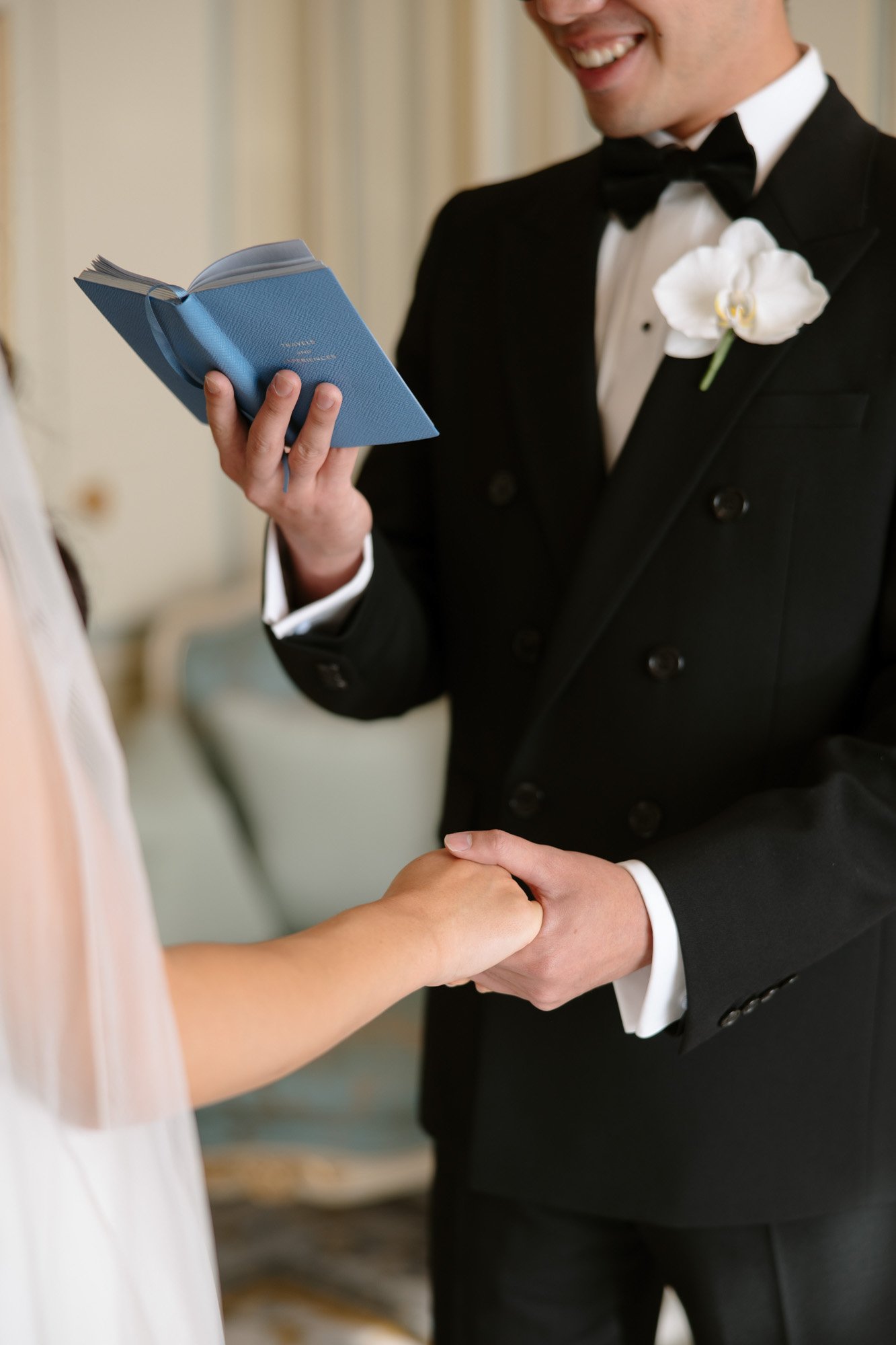 A man in a tuxedo reads from a blue book while holding hands with a person in a wedding dress.
