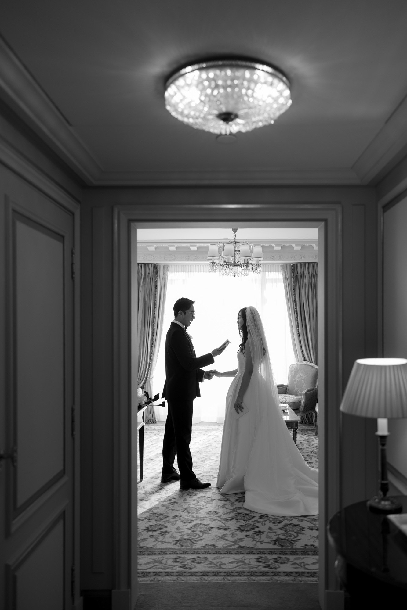 A bride and groom stand facing each other in a well-lit room, holding papers, with formal attire and elegant decor visible. Intimate Ritz Paris Elopement.