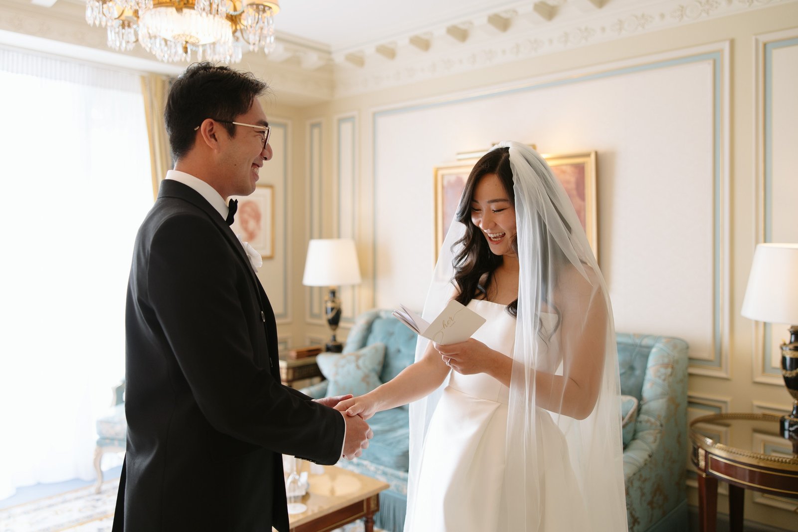 A bride in a white dress and veil reads from a card while holding hands with a groom in a black tuxedo in an elegant, well-lit room. Intimate Ritz Paris Elopement.