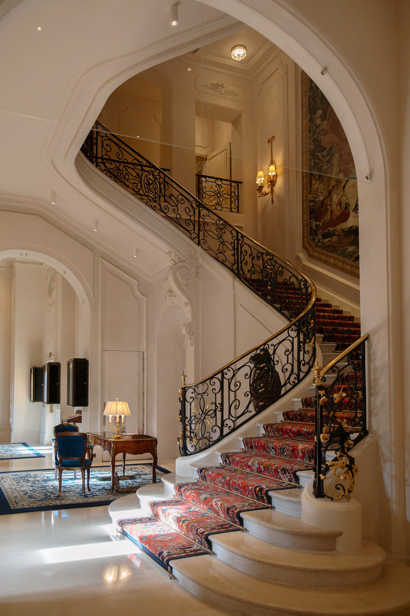 A grand staircase with ornate black and gold railing and red patterned carpet ascends in an elegant room with white walls, chandeliers, and classic furnishings. Intimate Ritz Paris Elopement.