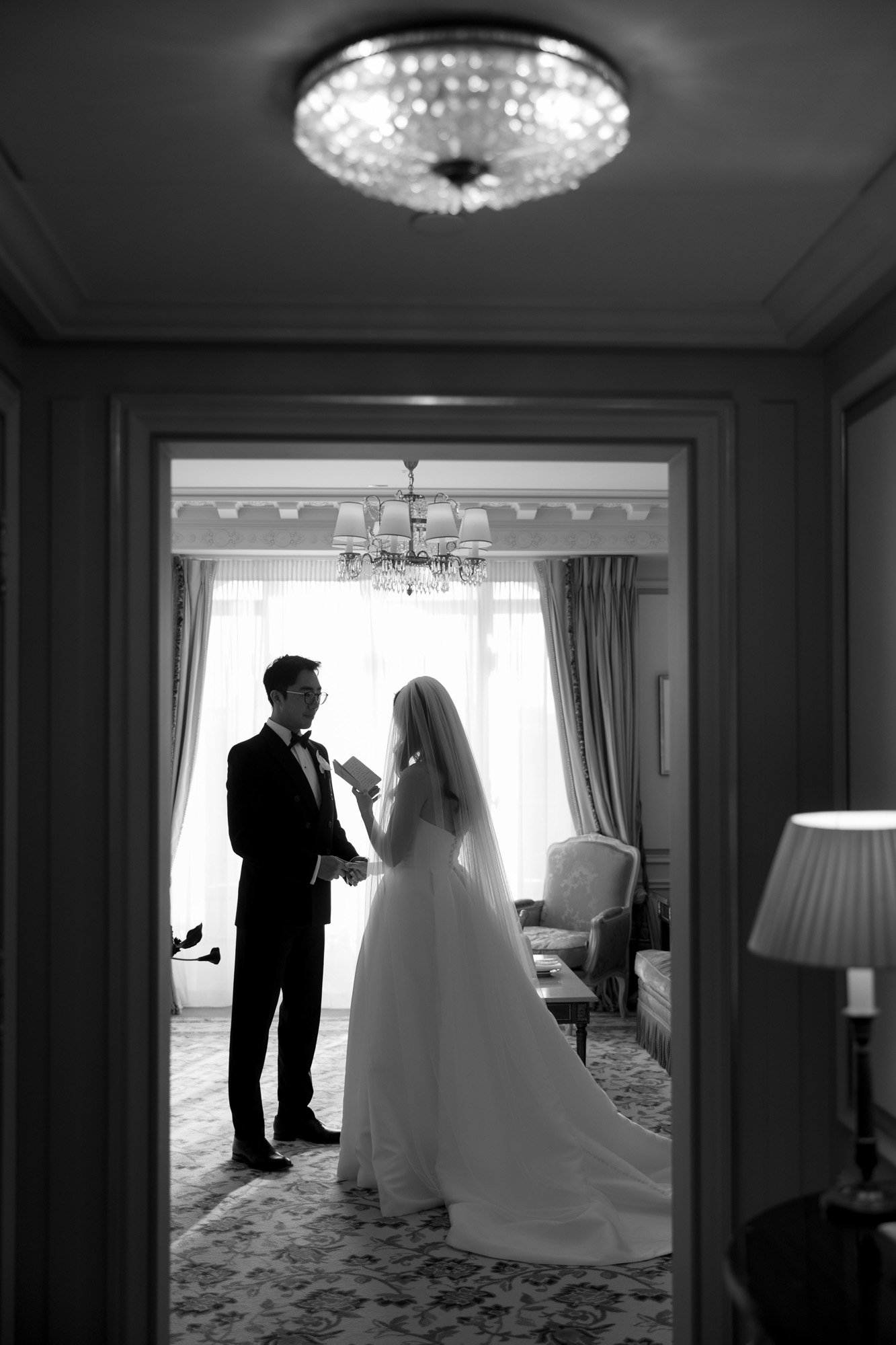 A bride and groom stand facing each other in a well-lit room, the bride holding a piece of paper, both dressed in formal wedding attire.
