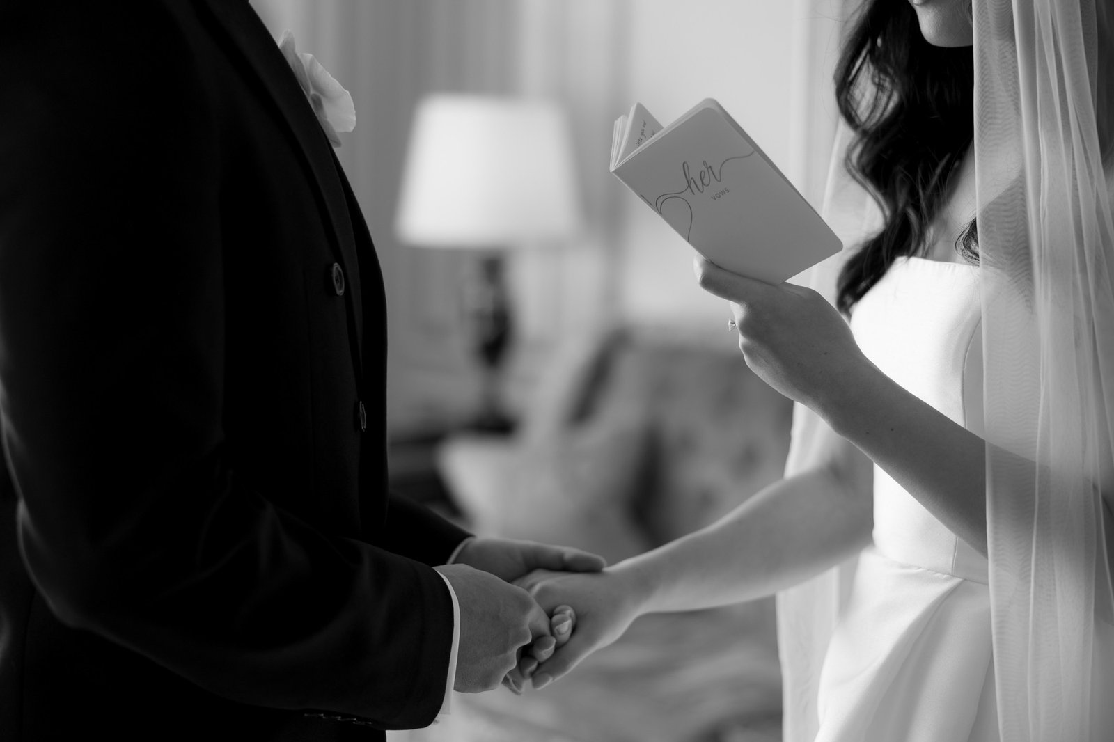 A bride in a white dress reads vows from a booklet while holding hands with a groom in a suit during a wedding ceremony.