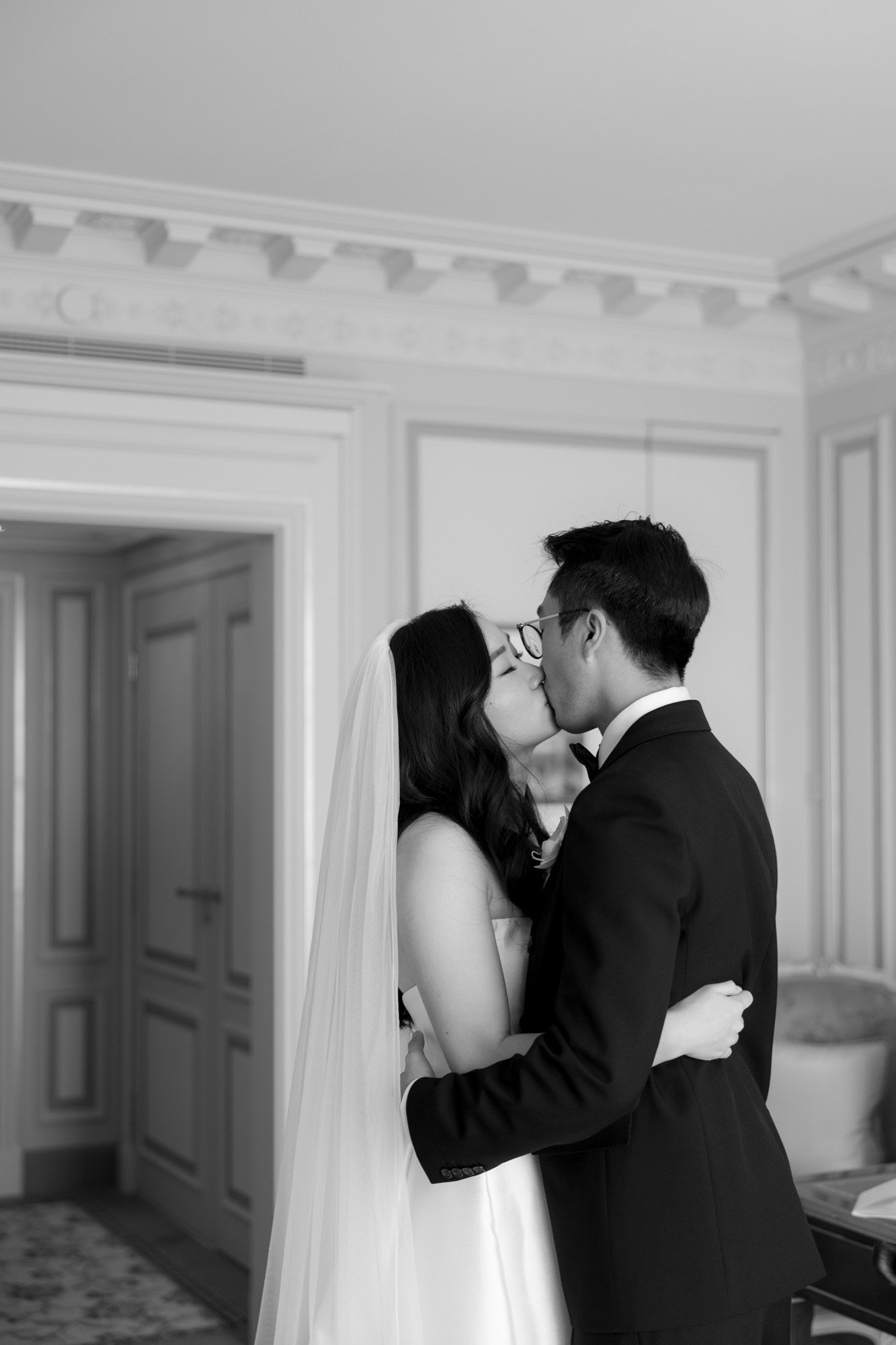 A bride and groom kiss in a well-lit room, with the bride wearing a veil and the groom in a suit. The scene is captured in black and white.