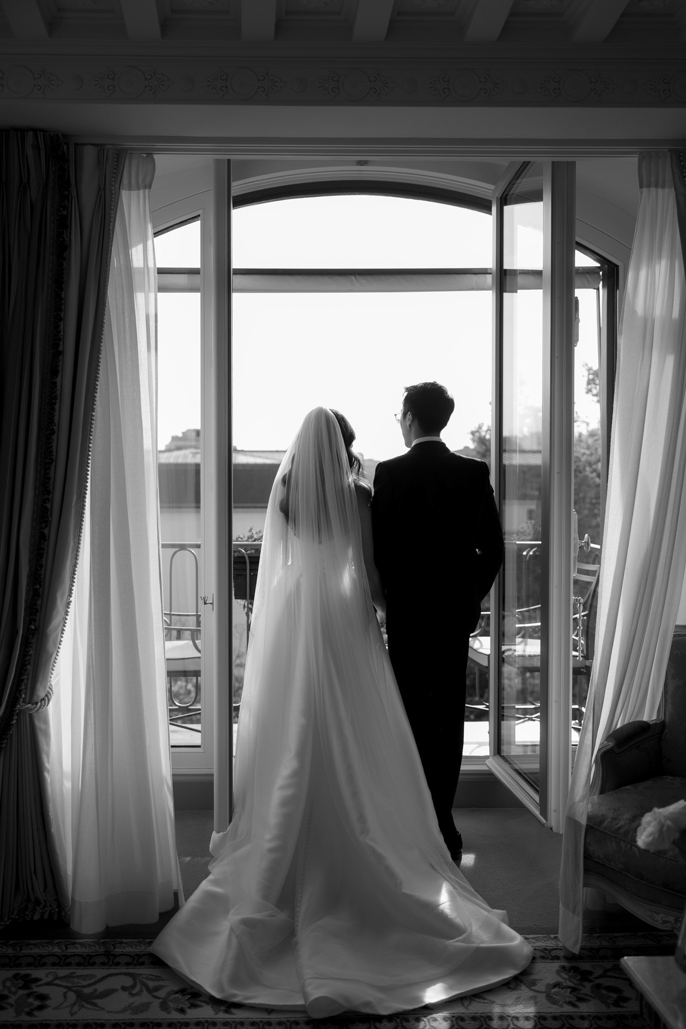 A bride and groom stand side by side, facing an open window with sheer curtains, looking outside. Intimate Ritz Paris Elopement.
