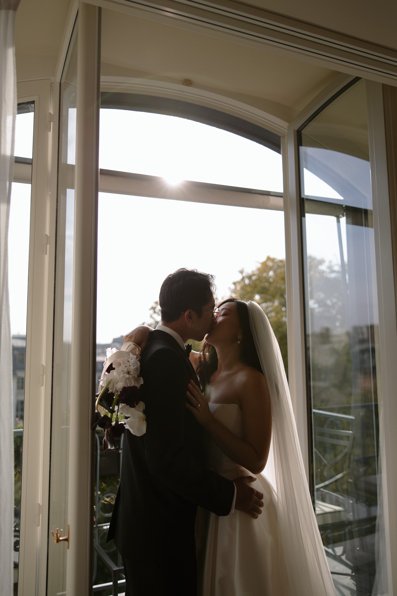 A bride and groom stand by an open balcony door, embracing and kissing in natural light, with the sun shining through the window behind them. Intimate Ritz Paris Elopement.