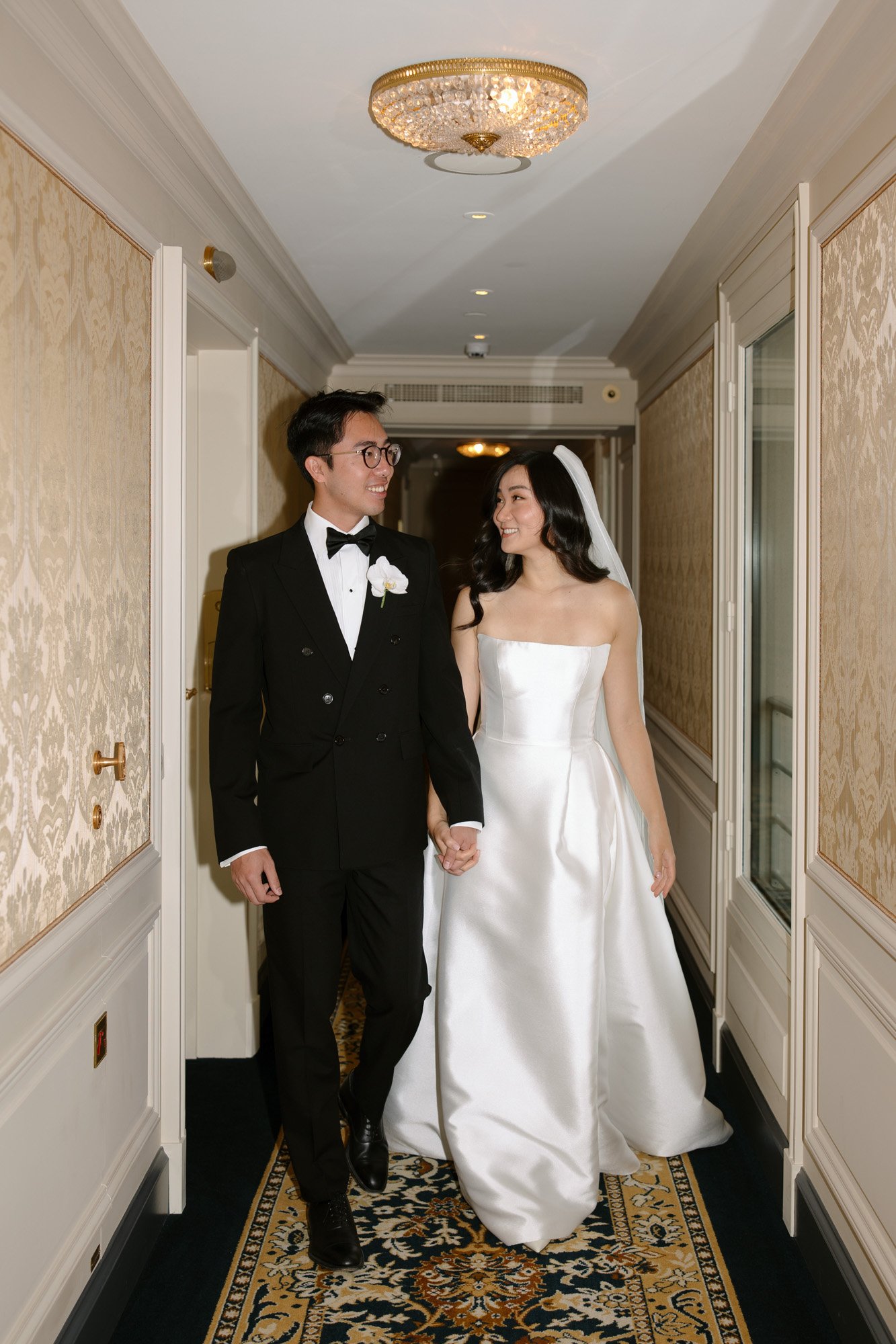 A bride in a white strapless gown and a groom in a black tuxedo walk hand in hand down a decorated hallway, smiling at each other. Intimate Ritz Paris Elopement.