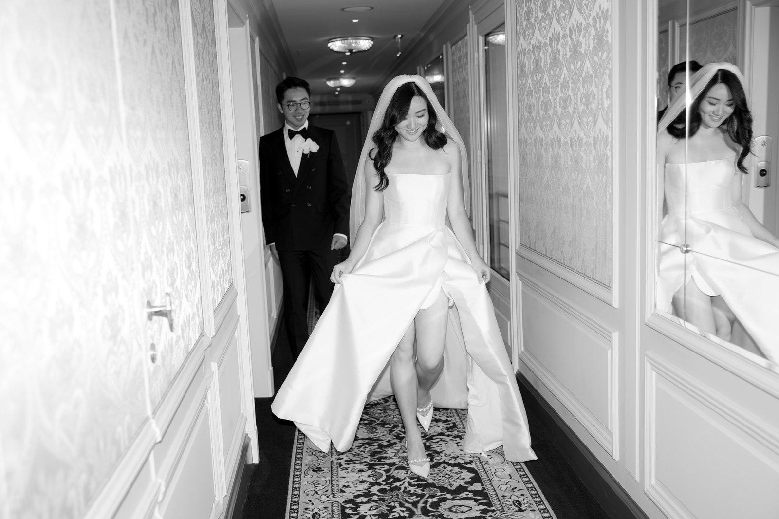 A bride in a strapless gown walks down a hallway, holding her dress, while a groom in a tuxedo follows behind; their reflections are visible in the wall mirrors. Intimate Ritz Paris Elopement.