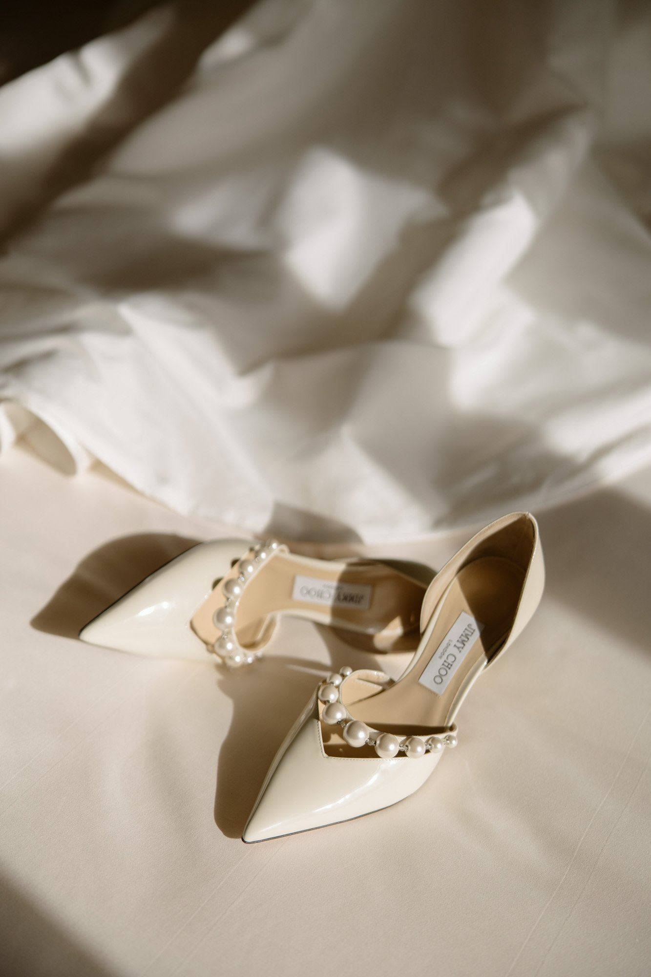 A pair of cream-colored pointed-toe heels with pearl embellishments rests on a light fabric surface under soft lighting. Intimate Ritz Paris Elopement.