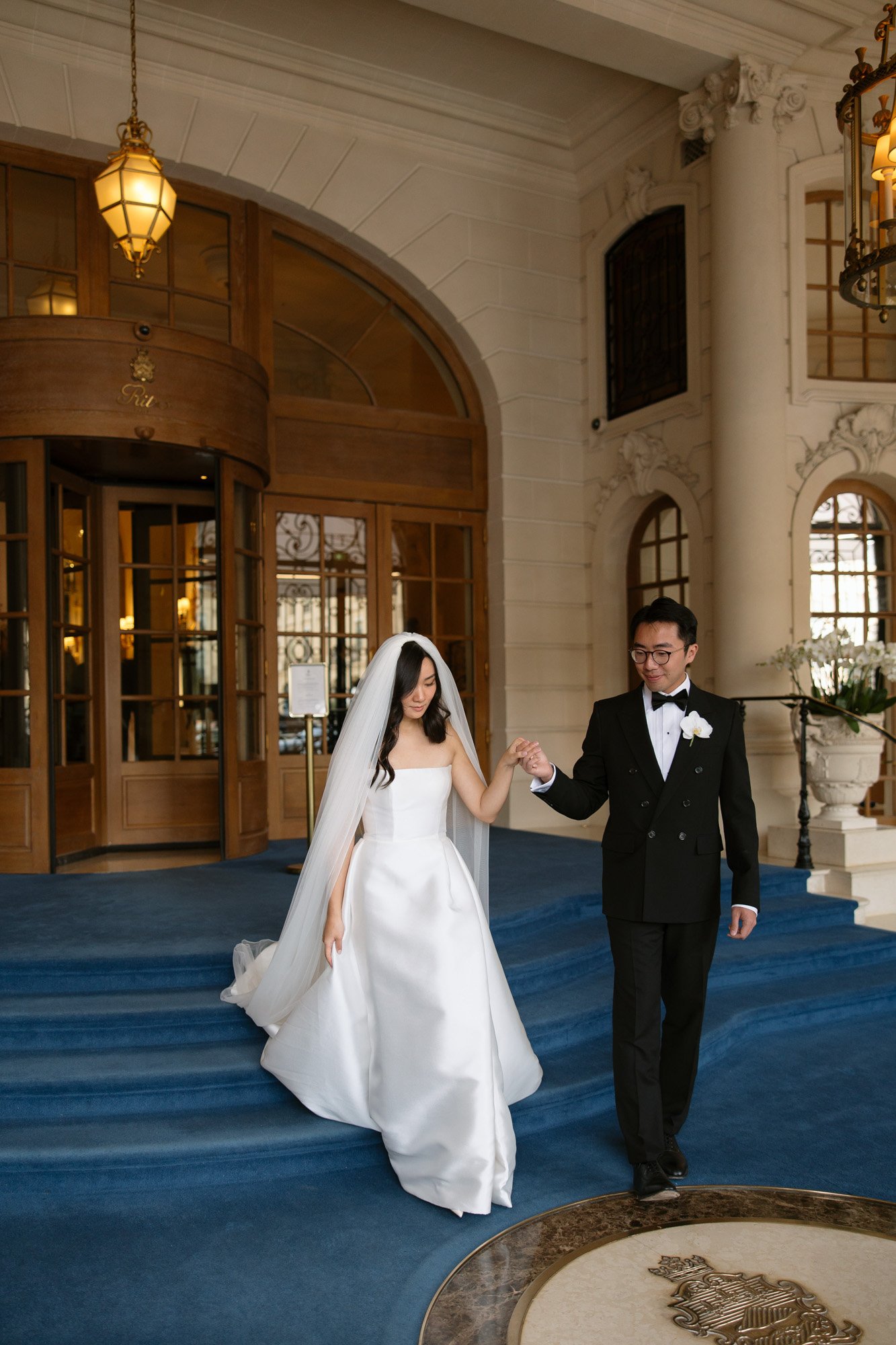 A bride and groom in formal attire walk down blue-carpeted steps inside an elegant building with columns and large wooden doors. Paris wedding photographer.