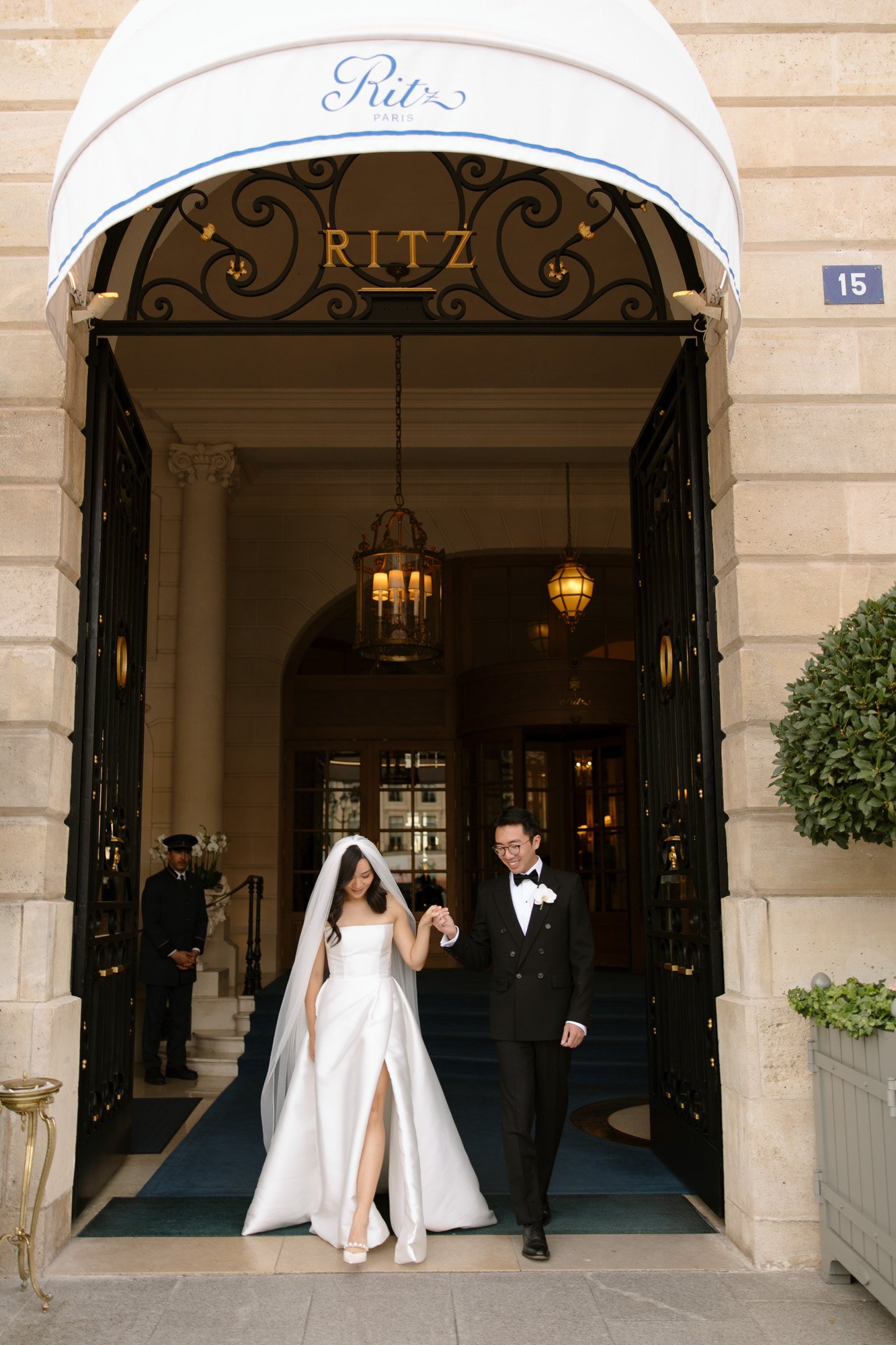 A bride and groom walk out of the Ritz Paris hotel entrance, with the bride in a white gown and veil and the groom in a black tuxedo.