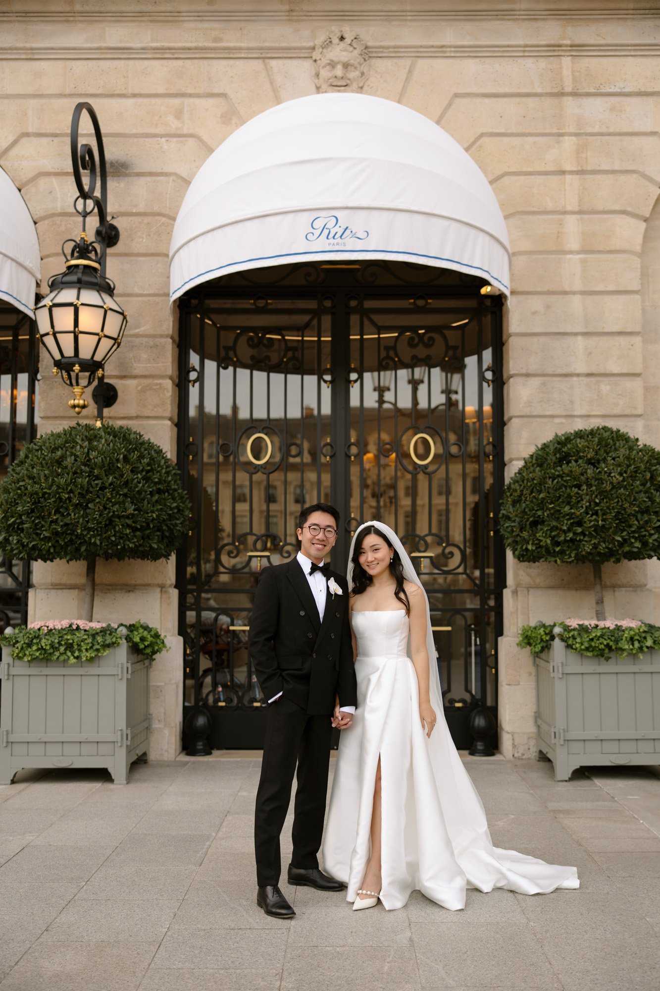 A bride and groom stand side by side in formal wedding attire in front of an ornate entrance with Ritz on the awning. Intimate Ritz Paris Elopement.