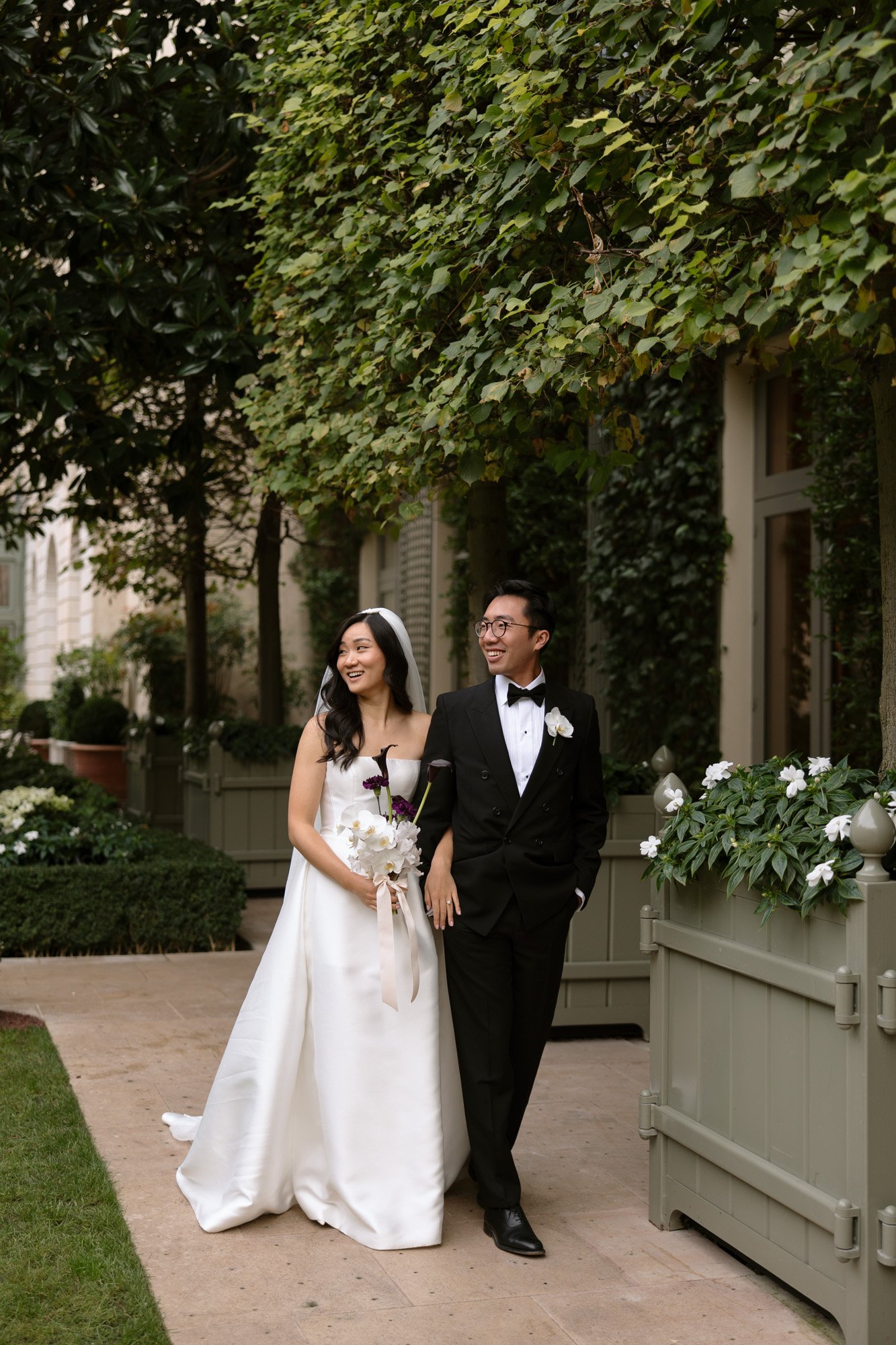 A bride in a white gown and a groom in a black tuxedo walk side by side outdoors, smiling and holding hands, surrounded by greenery. Intimate Ritz Paris Elopement.