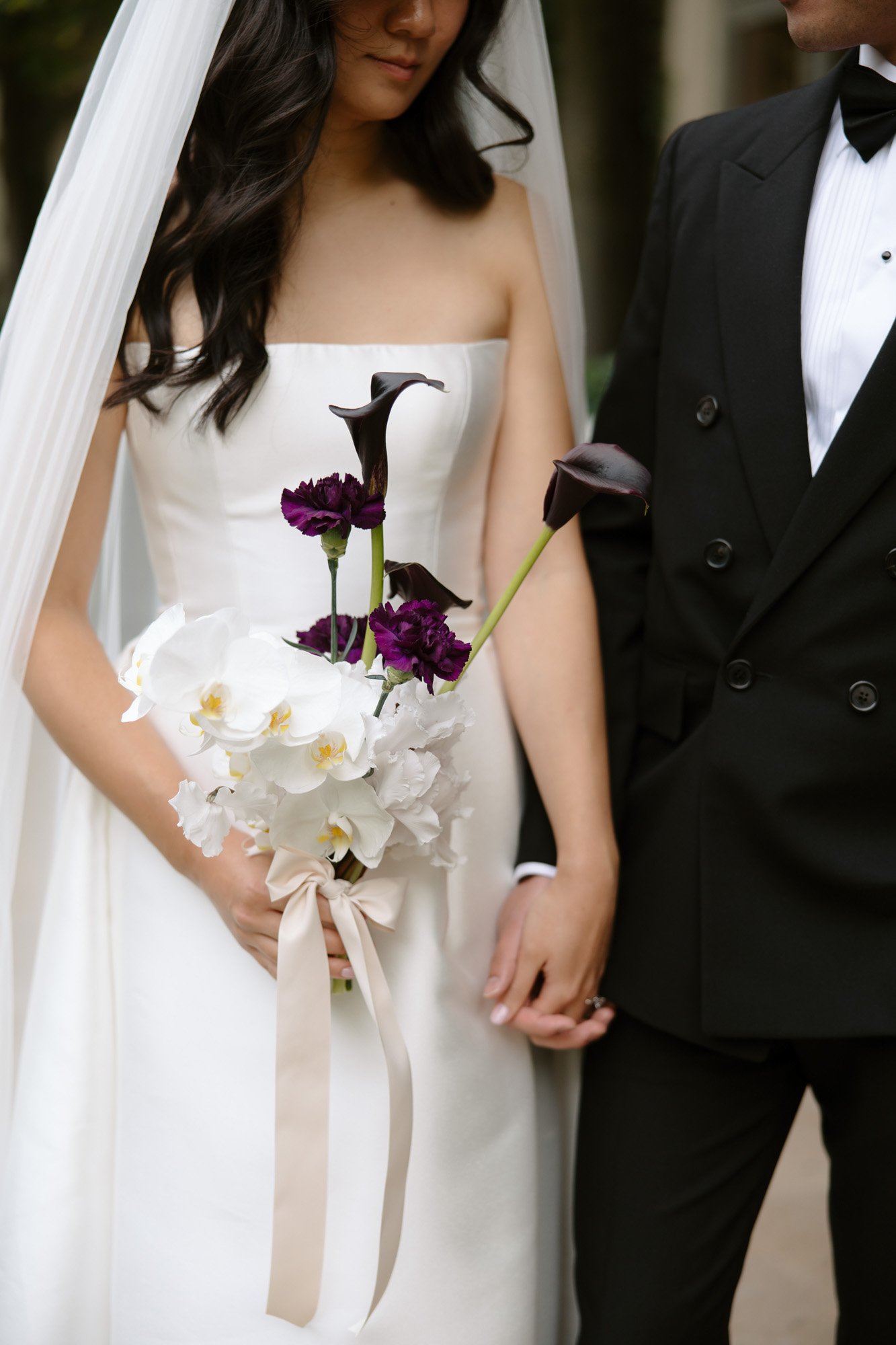A bride in a white dress holds a bouquet of white and dark purple flowers while standing and holding hands with a groom in a black tuxedo. Intimate Ritz Paris Elopement.