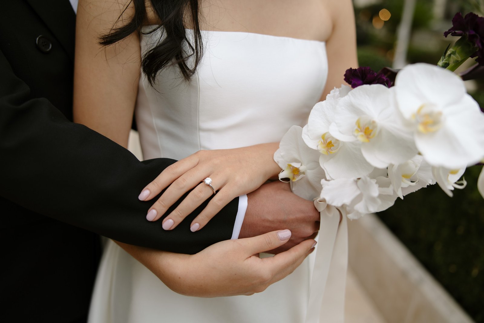 A bride in a white dress holds a bouquet of white orchids while a groom in a black suit embraces her from behind, showing her engagement ring.