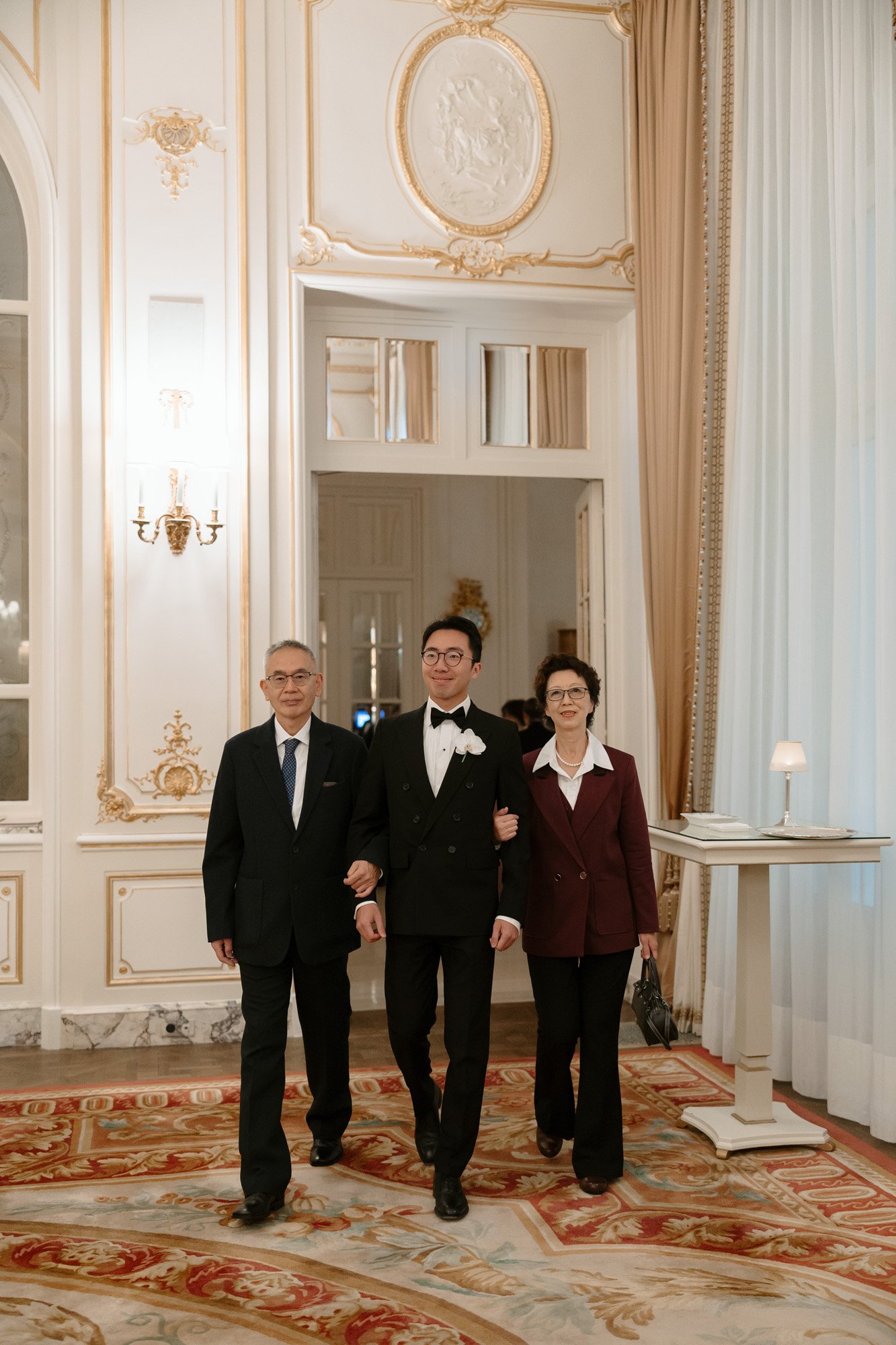 Three people in formal attire walk arm-in-arm through an elegant, ornate room with large windows, detailed molding, and a patterned carpet.