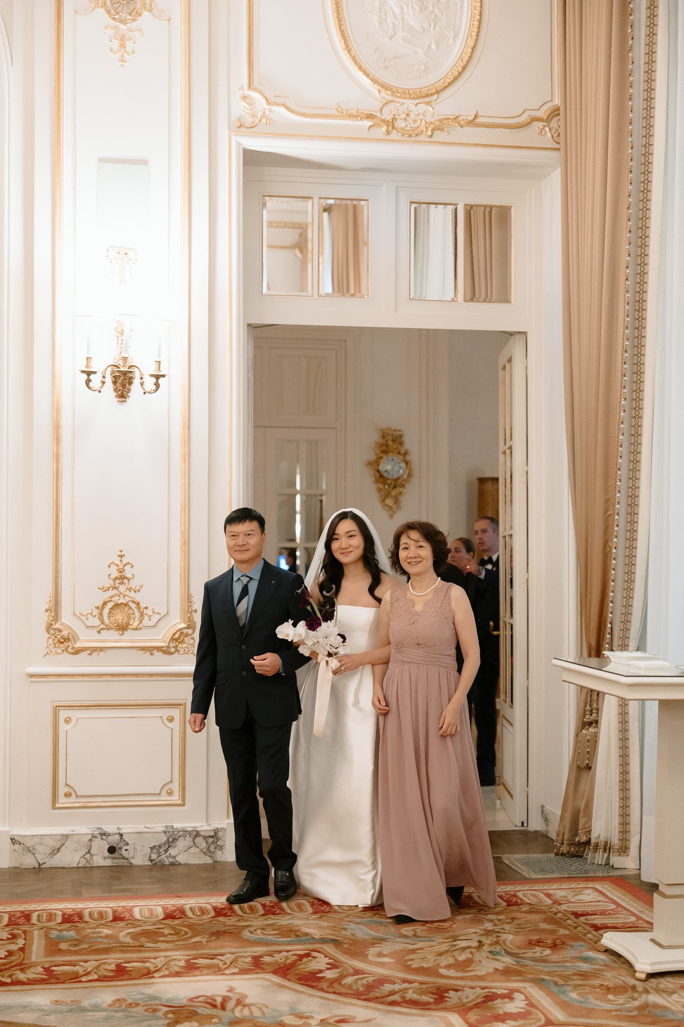 A bride in a white dress walks down the aisle flanked by two people in formal attire inside an ornately decorated room.
