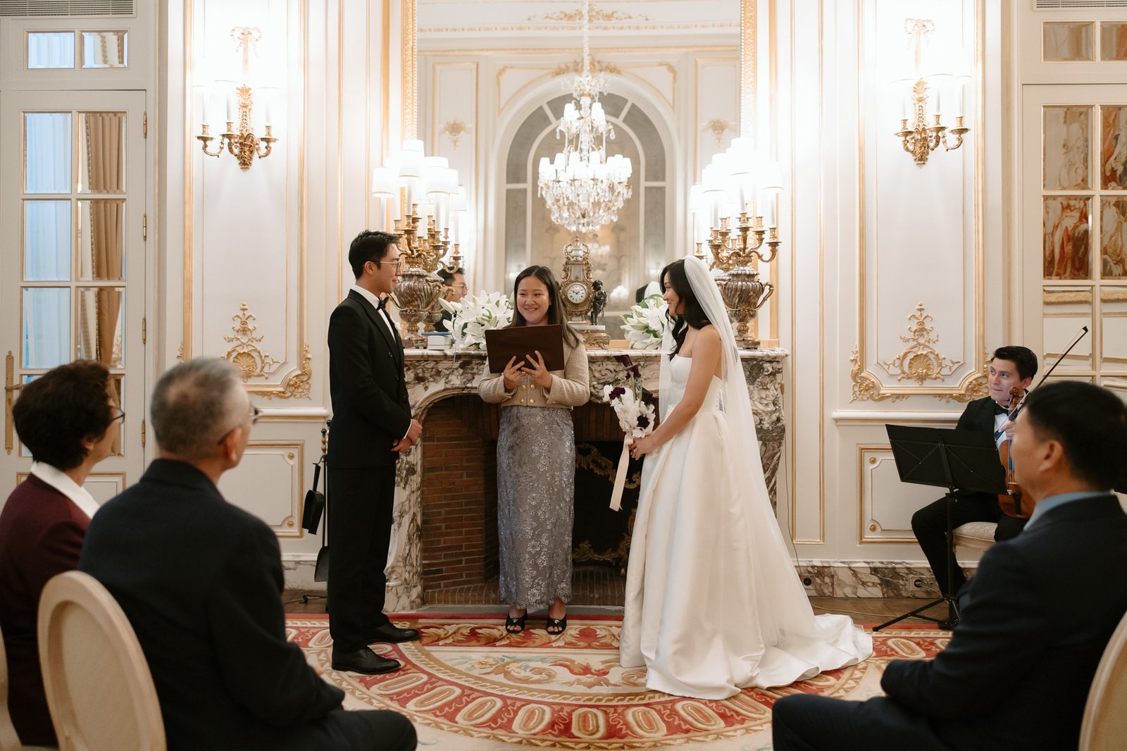 A bride and groom stand before an officiant during a wedding ceremony in an elegant, ornate room, with guests seated and musicians to the side. Intimate Ritz Paris Elopement.