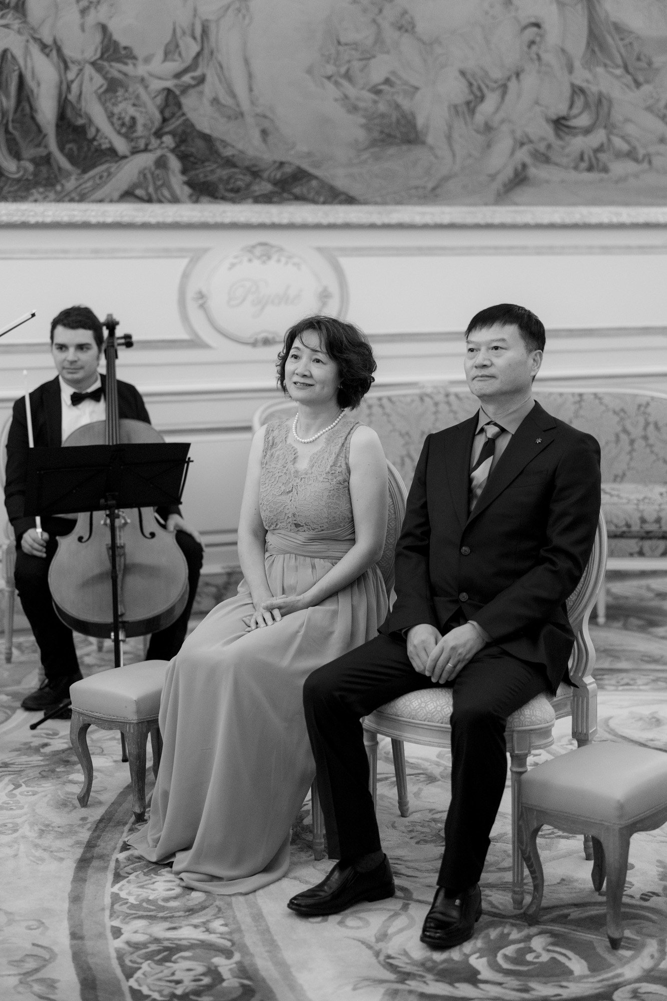 A woman in a formal dress and a man in a suit sit side by side on chairs in an ornate room, with a cellist seated behind them.