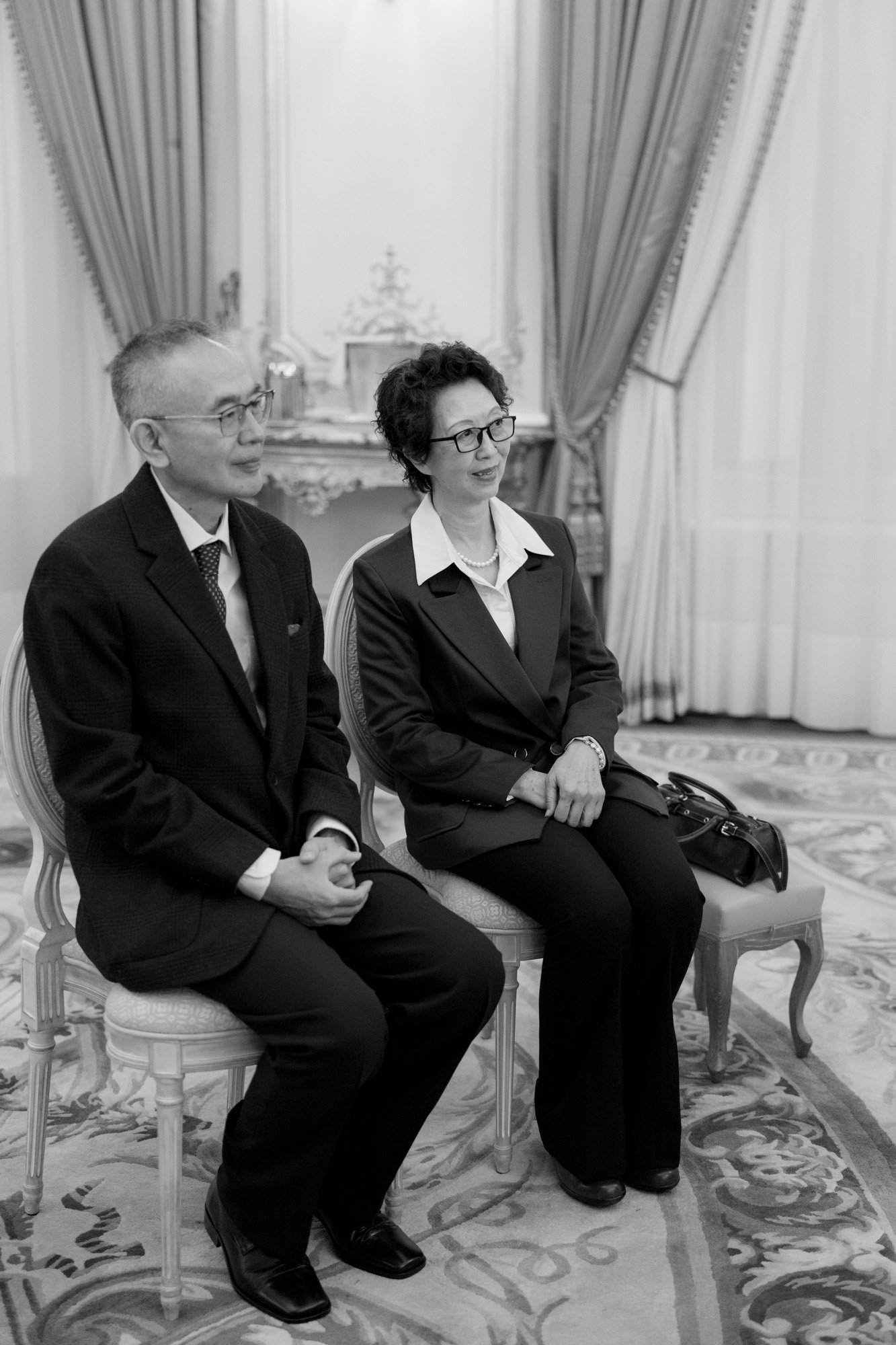 A man and a woman in formal attire sit side by side on chairs in an ornately decorated room with draped curtains and patterned carpet.
