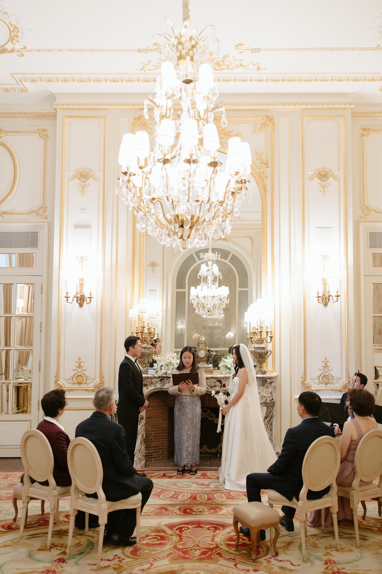 A wedding ceremony takes place indoors, with a couple standing before an officiant. Guests are seated around them in an ornate, chandelier-lit room with gold accents. Paris wedding photographer.
