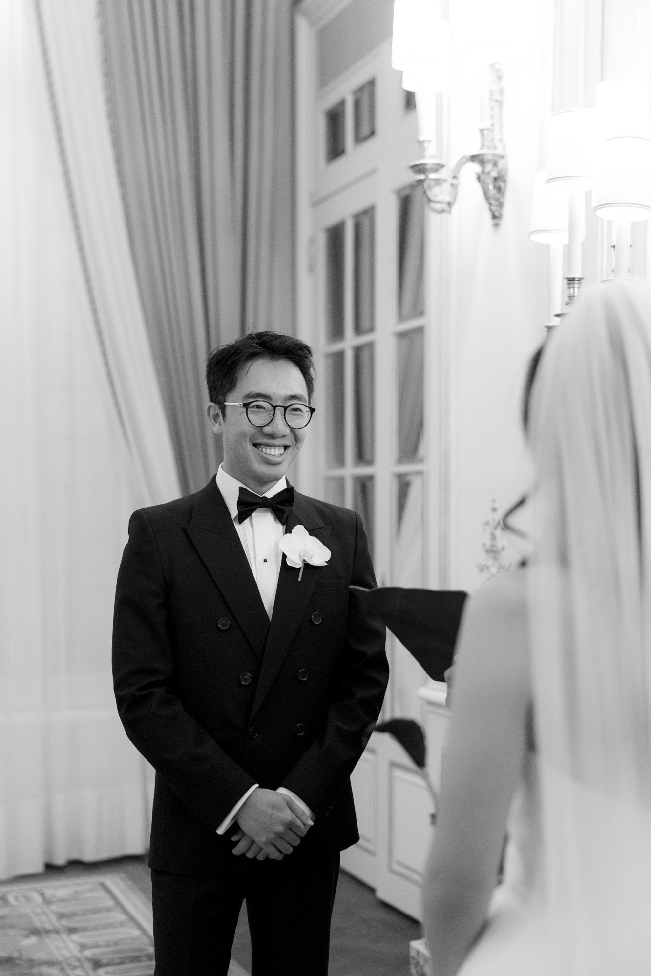 A man in a tuxedo smiles at a woman in a wedding dress and veil inside an elegantly decorated room.