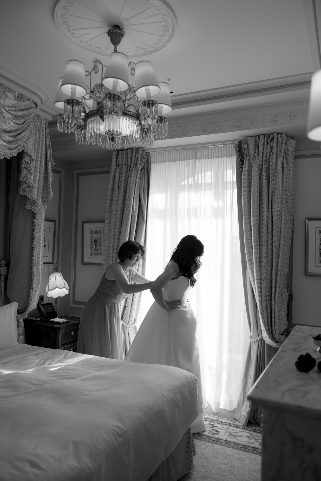 A woman helps another woman adjust her dress near a window in a well-decorated bedroom with elegant curtains and a chandelier.