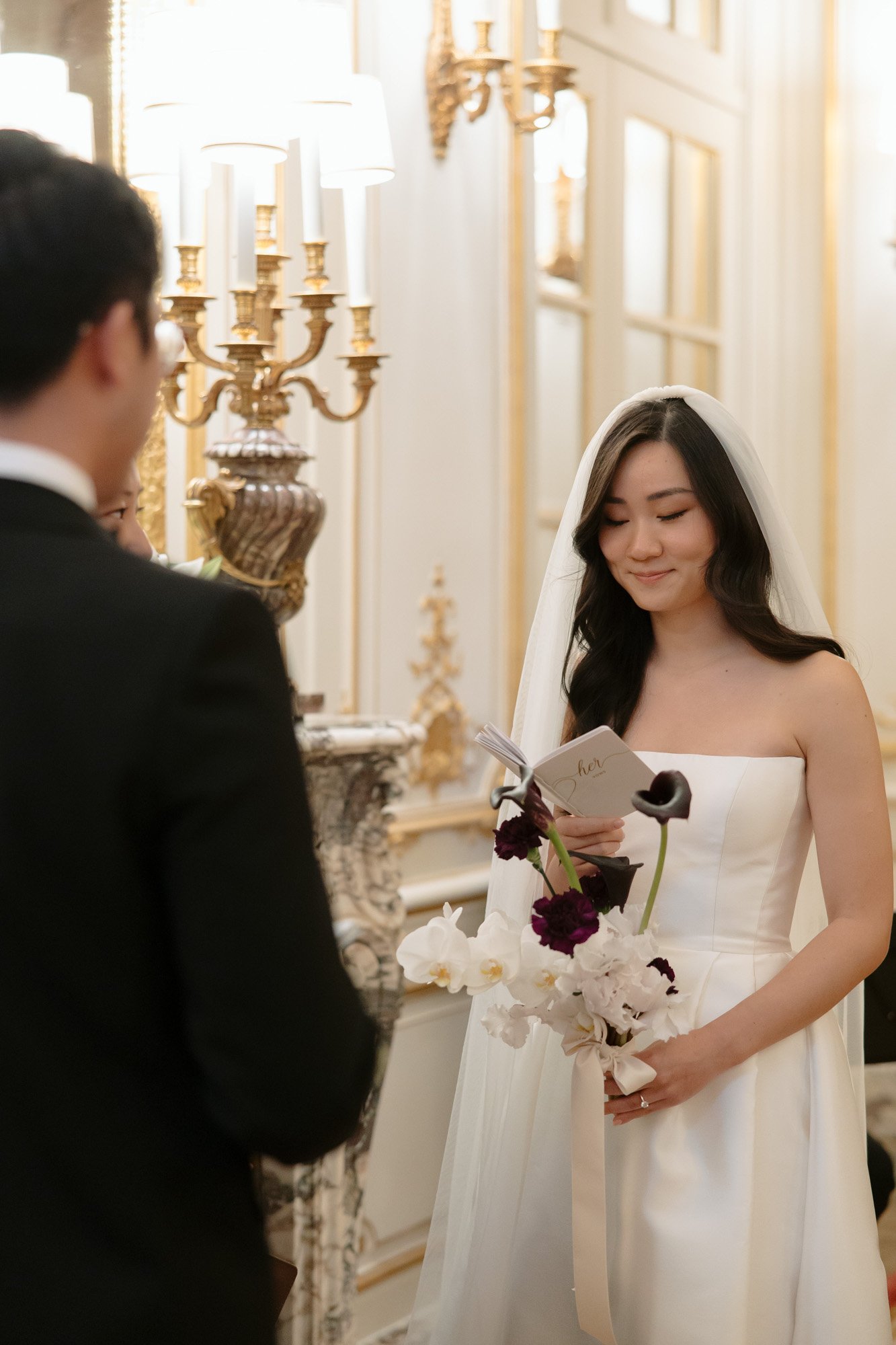 A bride in a white dress and veil holds a bouquet and reads from a card, standing opposite a groom in a formal indoor setting. Intimate Ritz Paris Elopement.