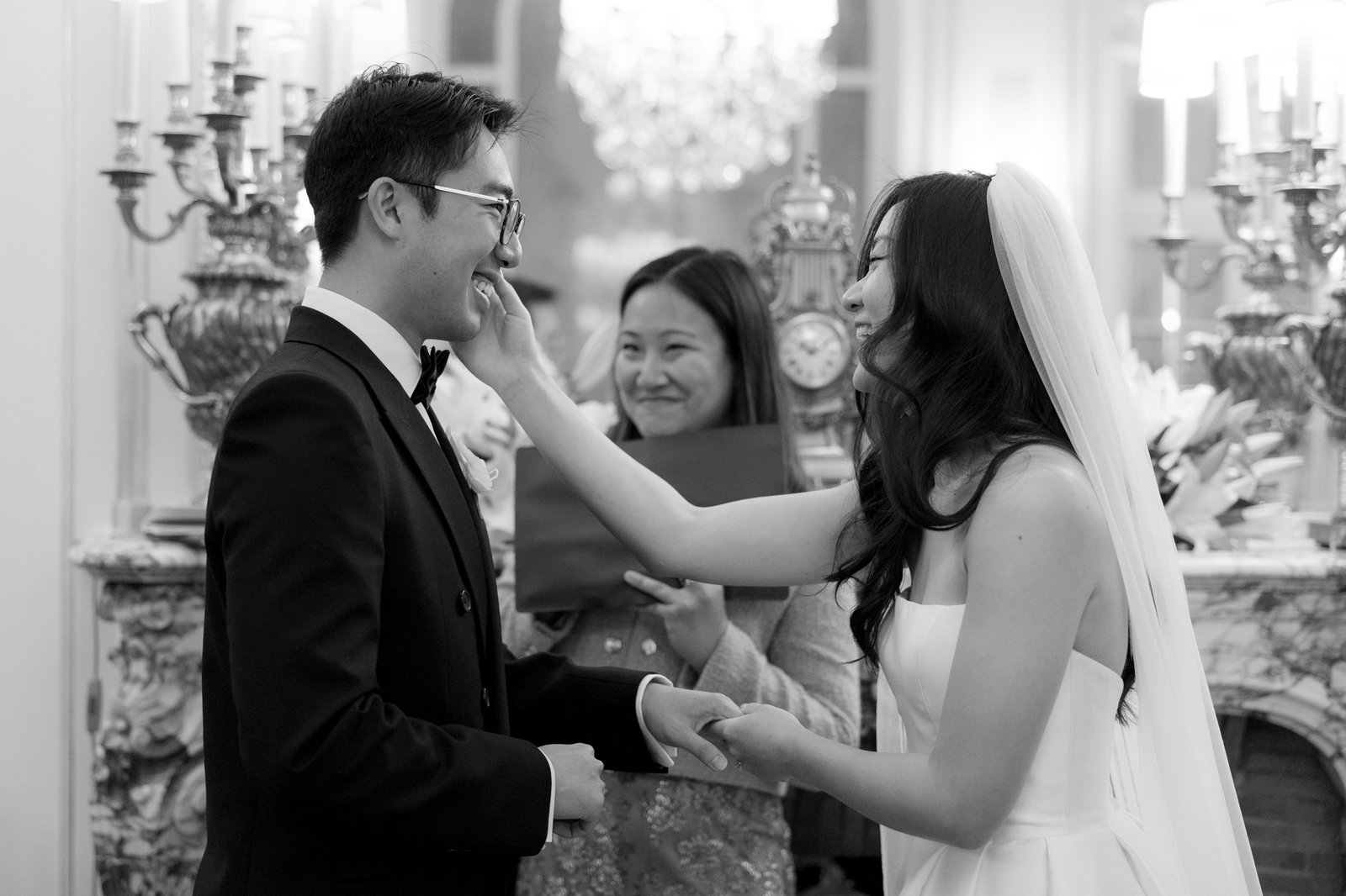 A bride and groom stand holding hands, smiling as the bride gently touches the grooms face during a wedding ceremony. An officiant stands behind them, also smiling. Intimate Ritz Paris Elopement.