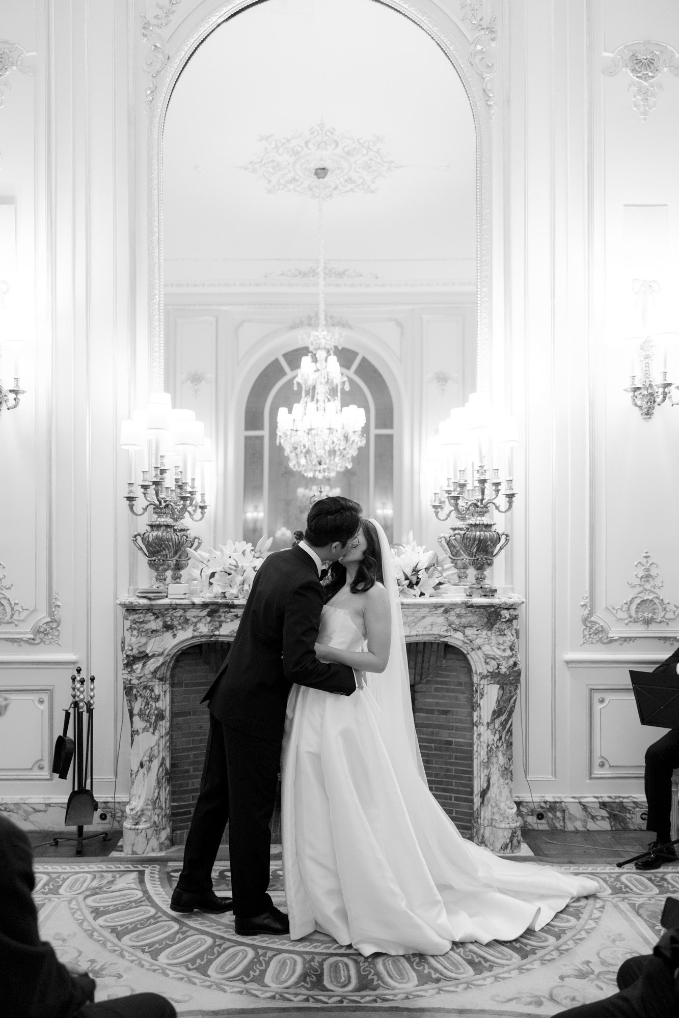 A bride and groom share a kiss at the altar in an ornate, elegant room with a large mirror, chandelier, and decorative fireplace. Intimate Ritz Paris Elopement.