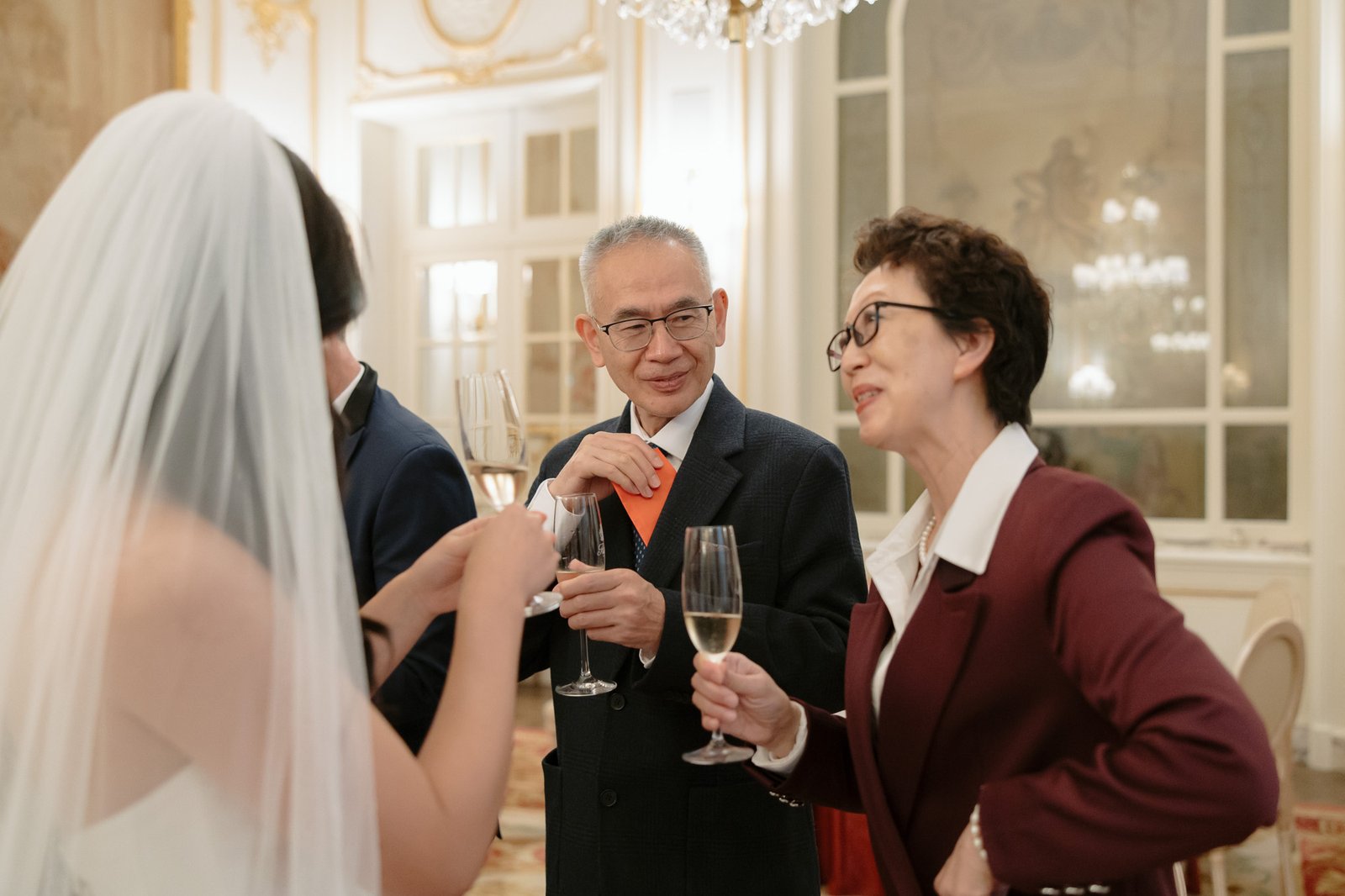 Four people in formal attire stand in an elegant room, holding champagne glasses and conversing; one is wearing a wedding dress with a veil.