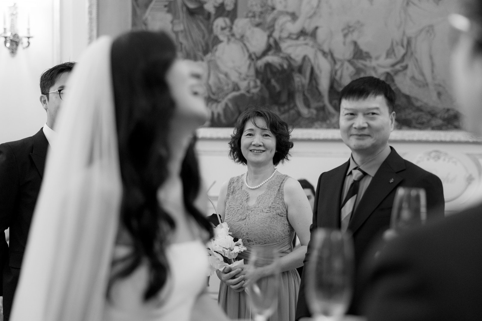A bride in the foreground is out of focus, while two guests, a woman in a lace dress and a man in a suit, stand smiling in the background at a formal event.
