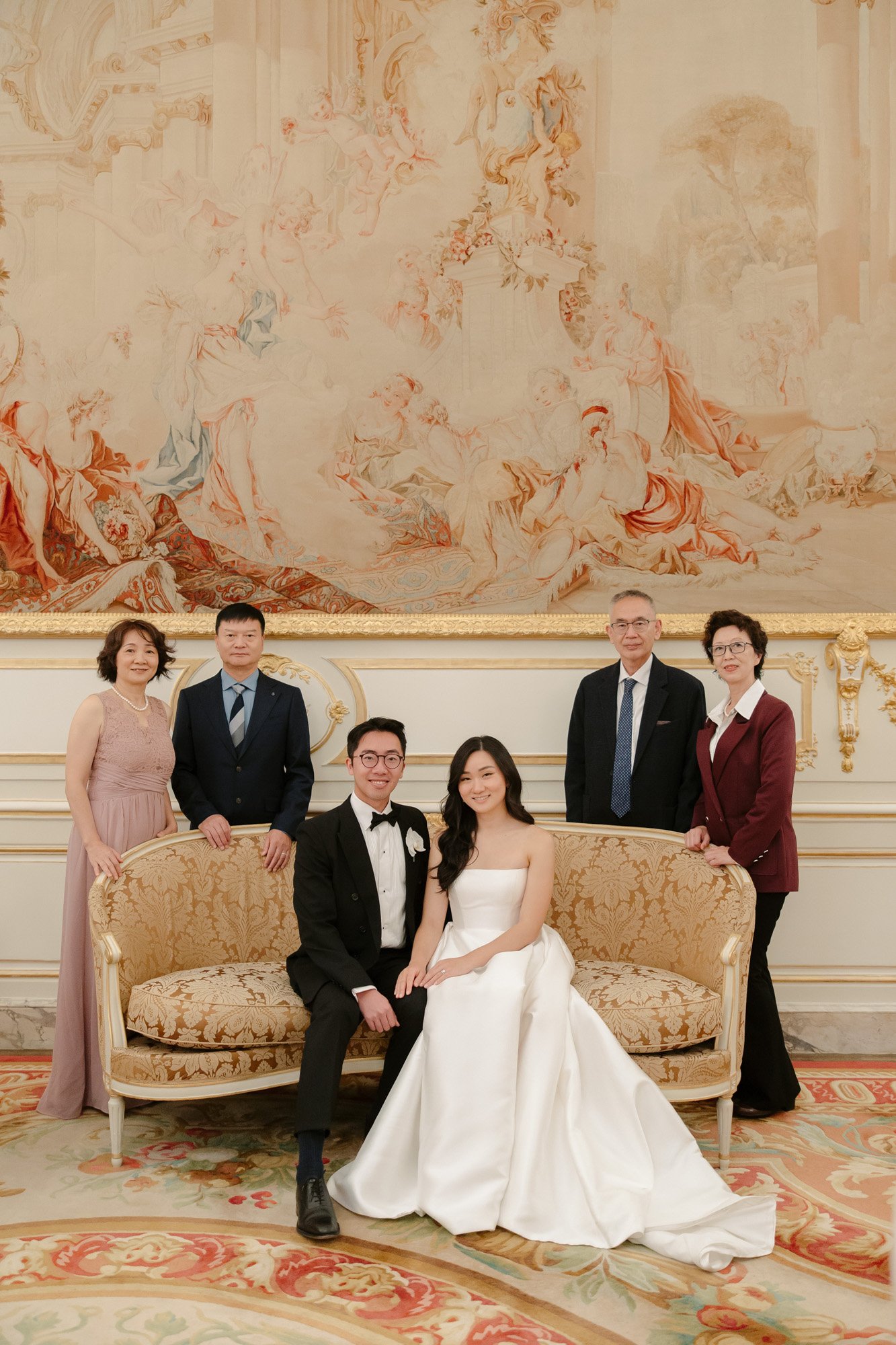 A bride and groom sit on a sofa with four adults standing behind them in an elegant, ornate room with a large tapestry. Intimate Ritz Paris Elopement.