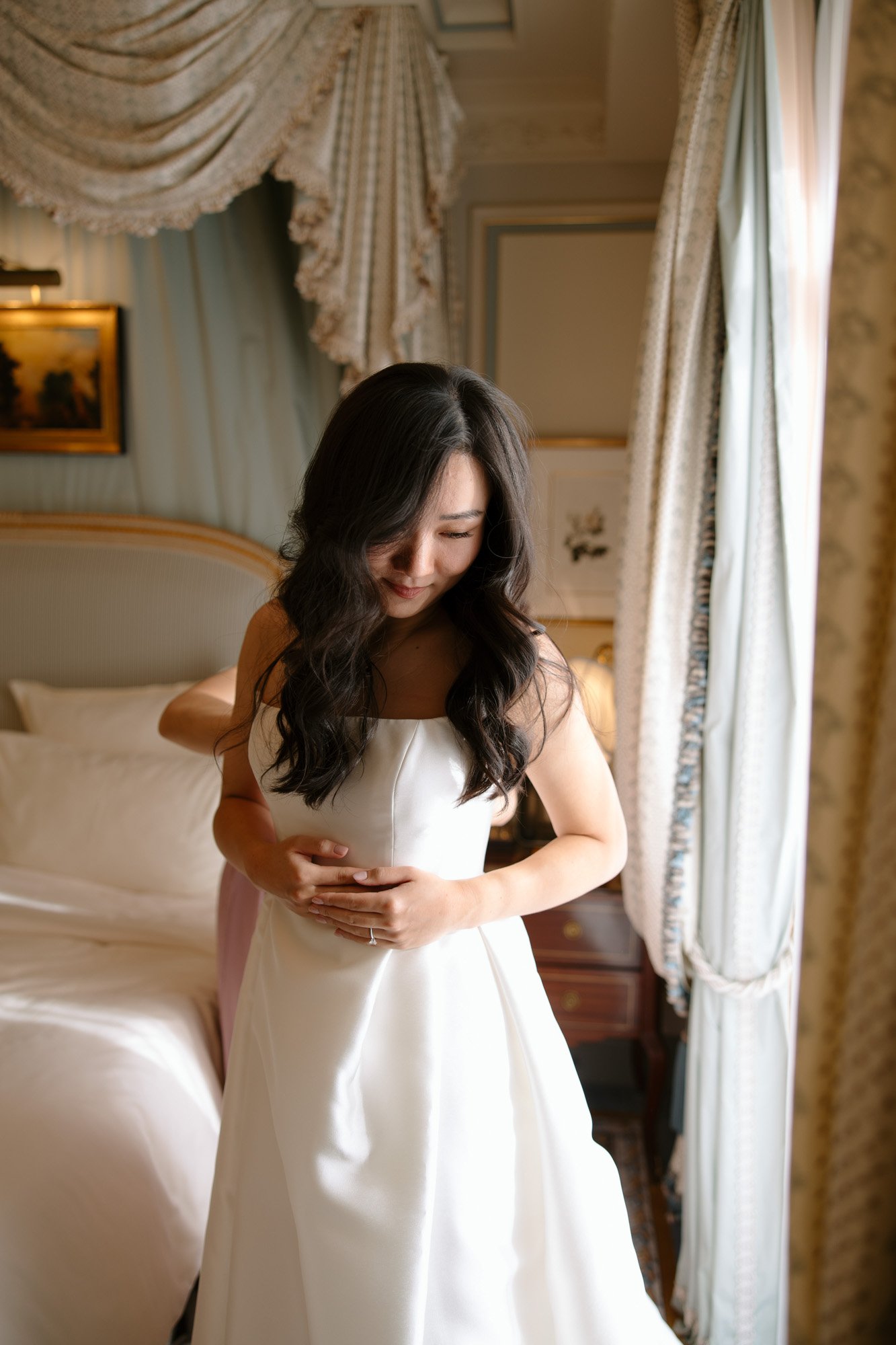A woman in a white dress stands in a softly lit room by a bed, with someone behind her adjusting the dress.