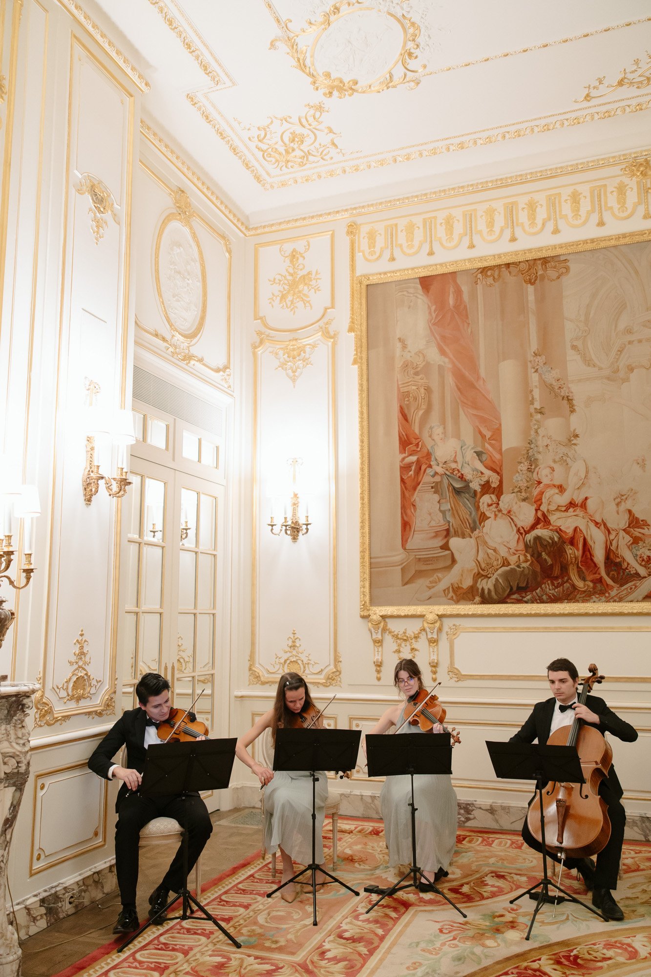 A string quartet performs in an ornate, gold-accented room with a large wall tapestry and classical decor.