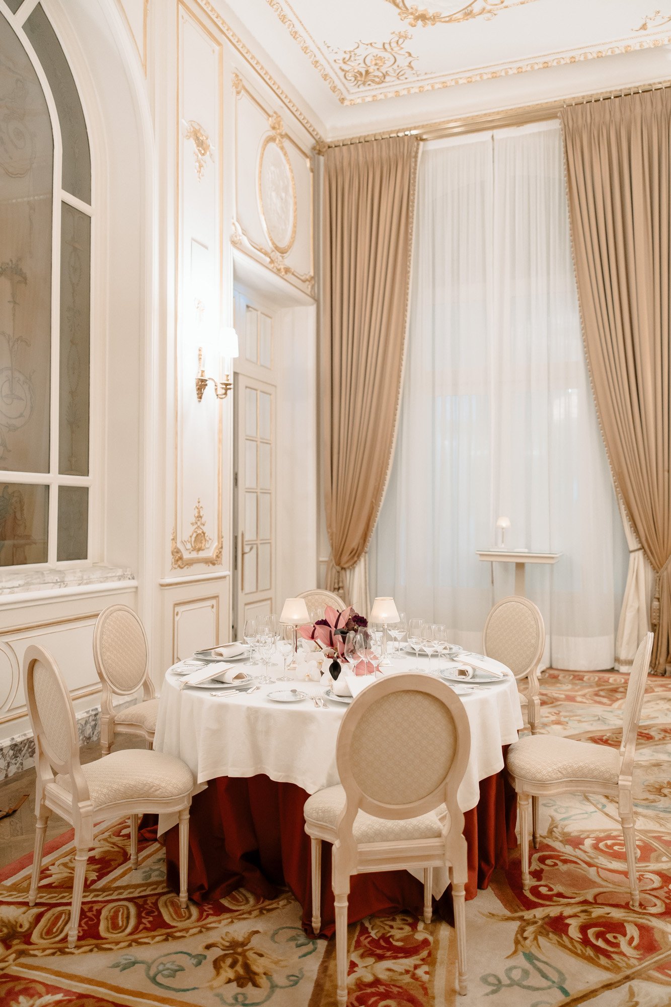 A round dining table with four chairs is set with glassware and napkins in an elegant, ornately decorated room with tall curtains and white walls. Intimate Ritz Paris Elopement.