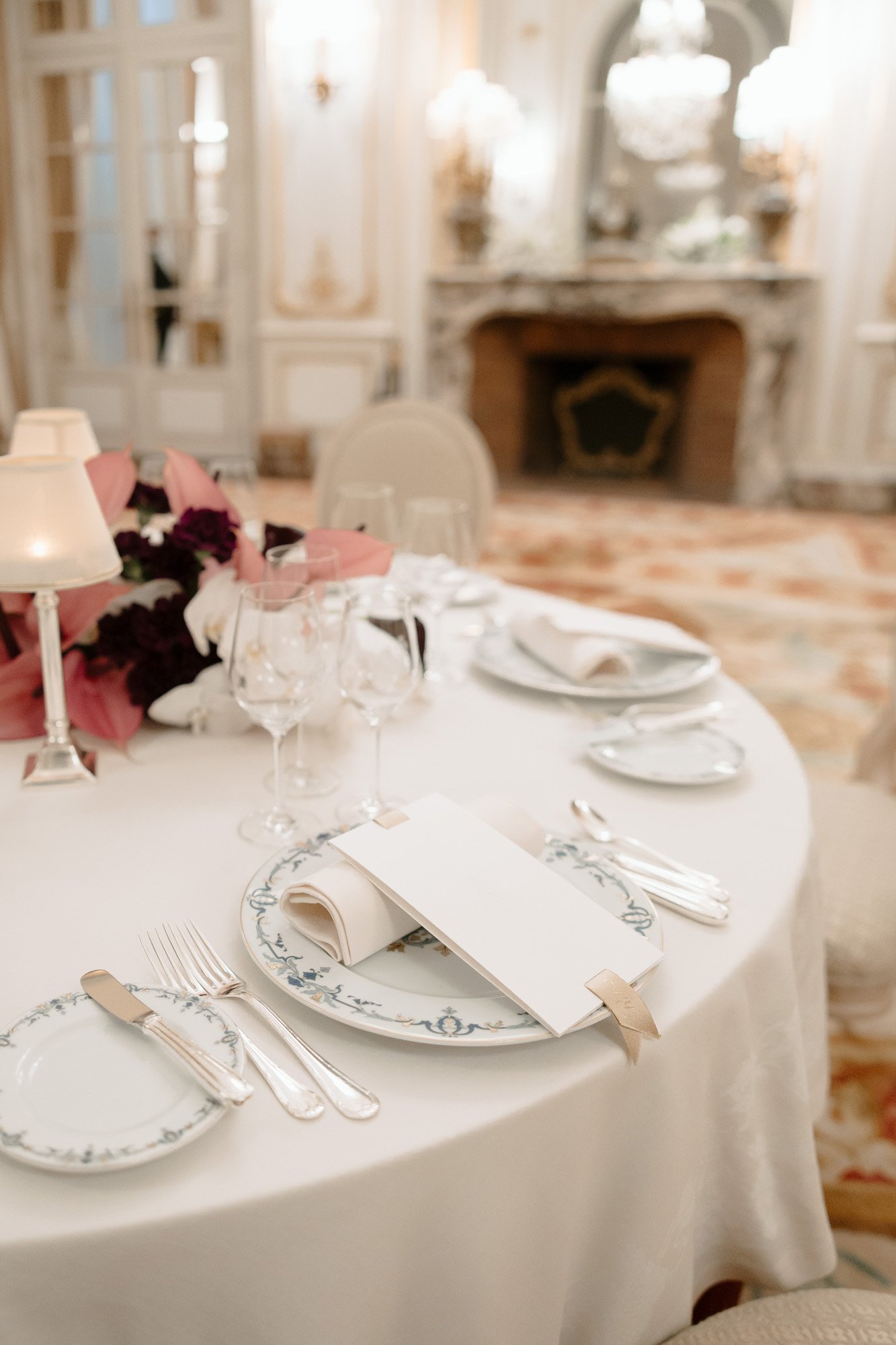 Elegant table setting with white and blue patterned plates, cutlery, wine glasses, a floral centerpiece, and a blank menu card in a formal dining room.