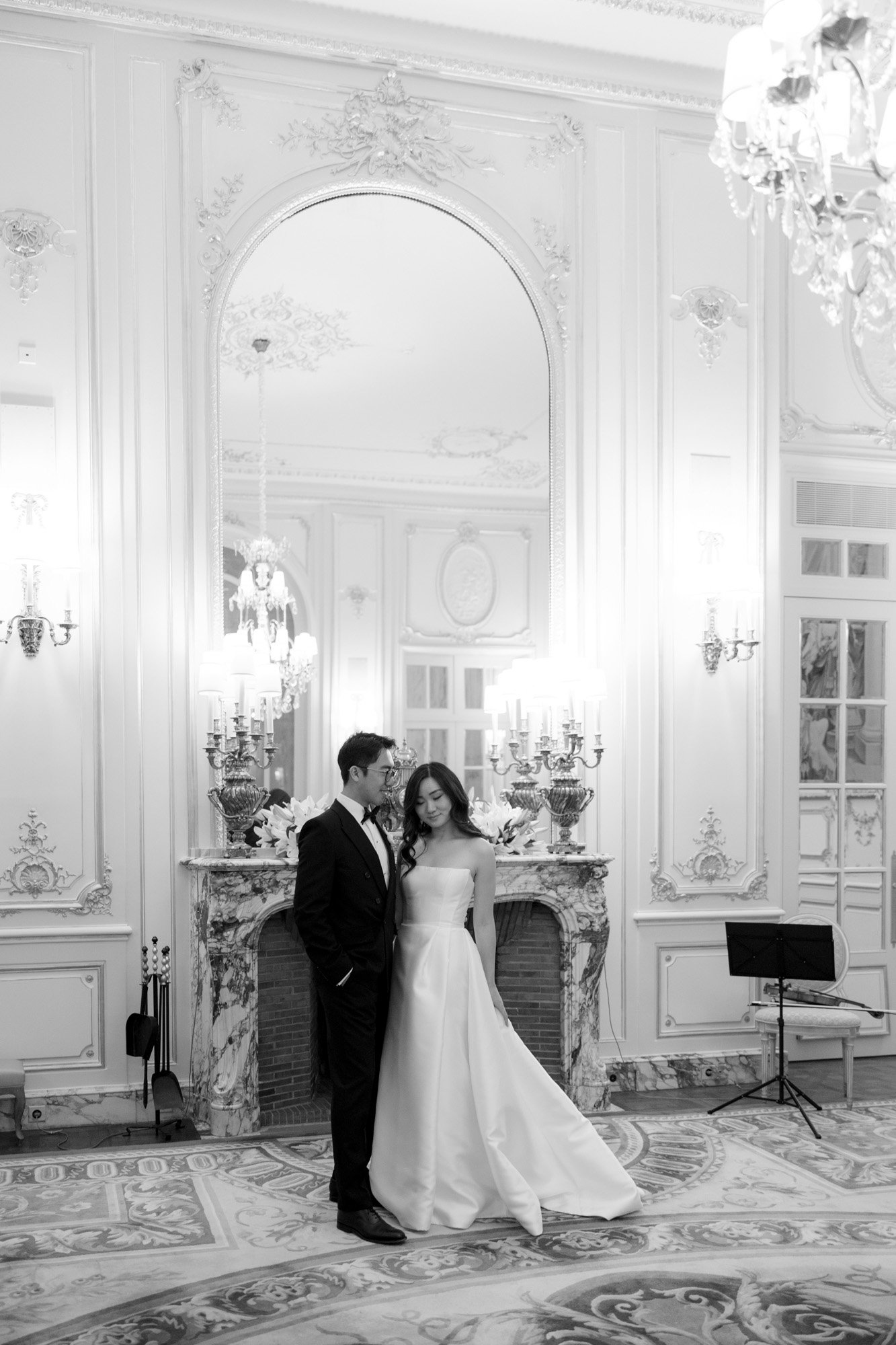 A bride and groom stand together in an ornate, elegant room with tall mirrors, chandeliers, and a decorated fireplace. Intimate Ritz Paris Elopement.