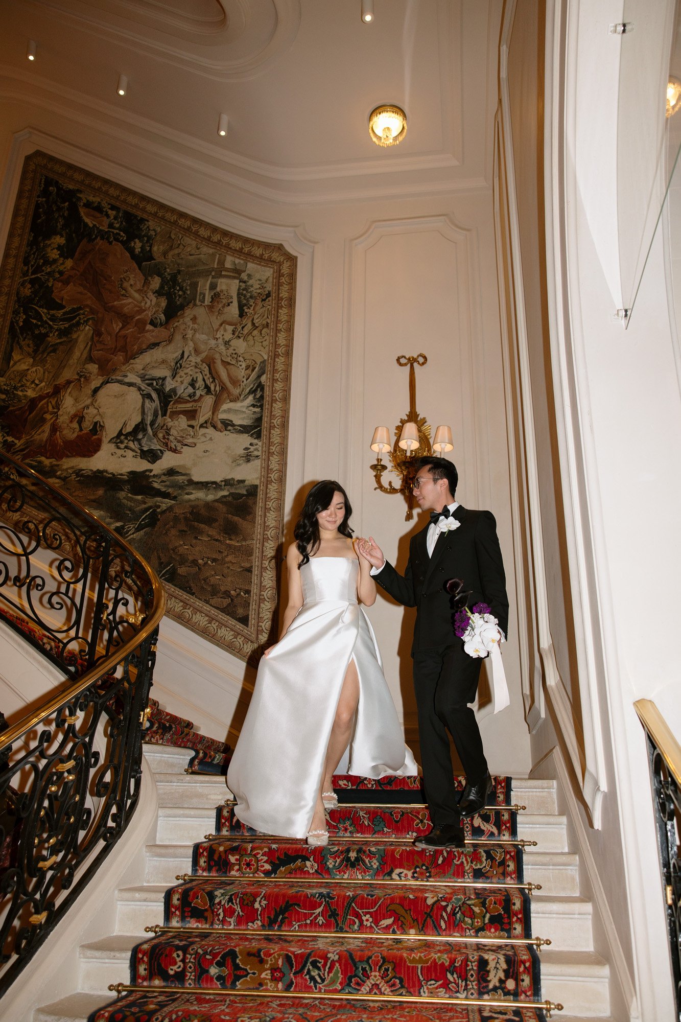 A couple in formal attire descends a carpeted staircase in an ornate interior with a large tapestry and decorative lighting on the wall. Intimate Ritz Paris Elopement.