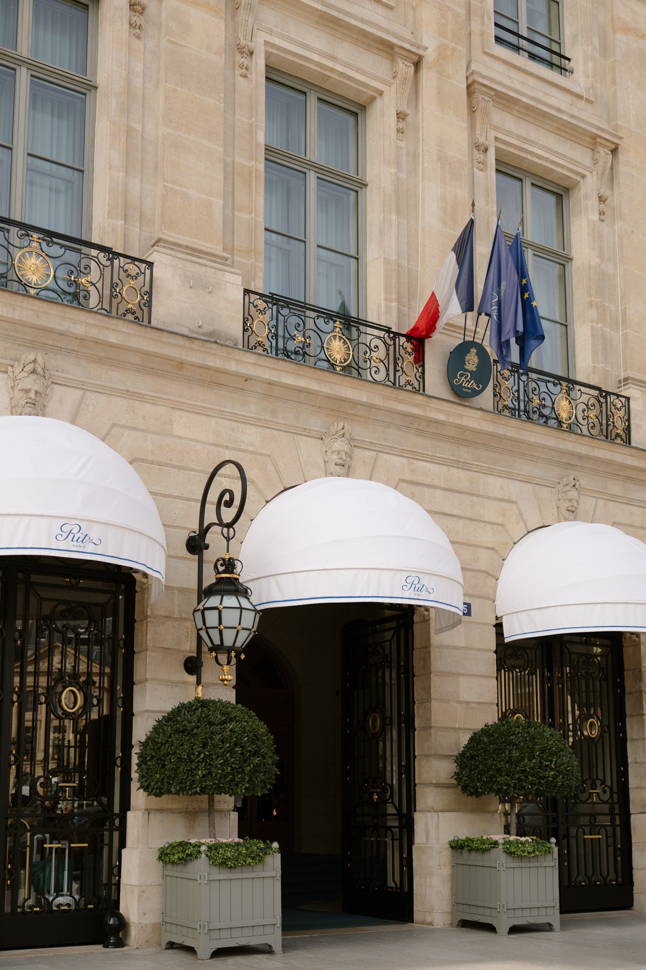 Elegant building entrance with ornate iron gates, two potted trees, white awnings labeled Ritz, and French and European Union flags above. Intimate Ritz Paris Elopement.