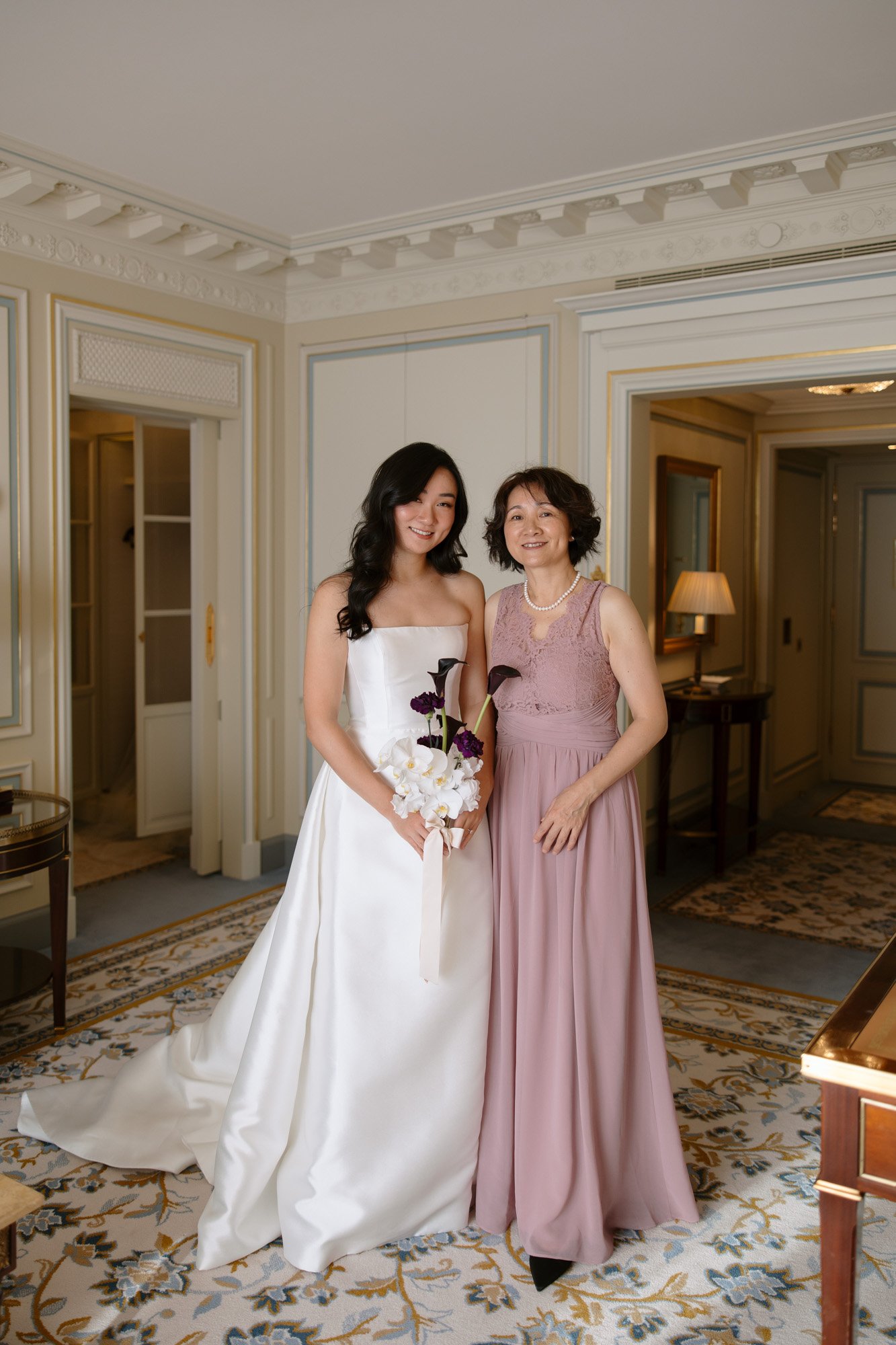 A bride in a white gown holding a bouquet stands next to a woman in a mauve dress, both smiling in a well-decorated room.