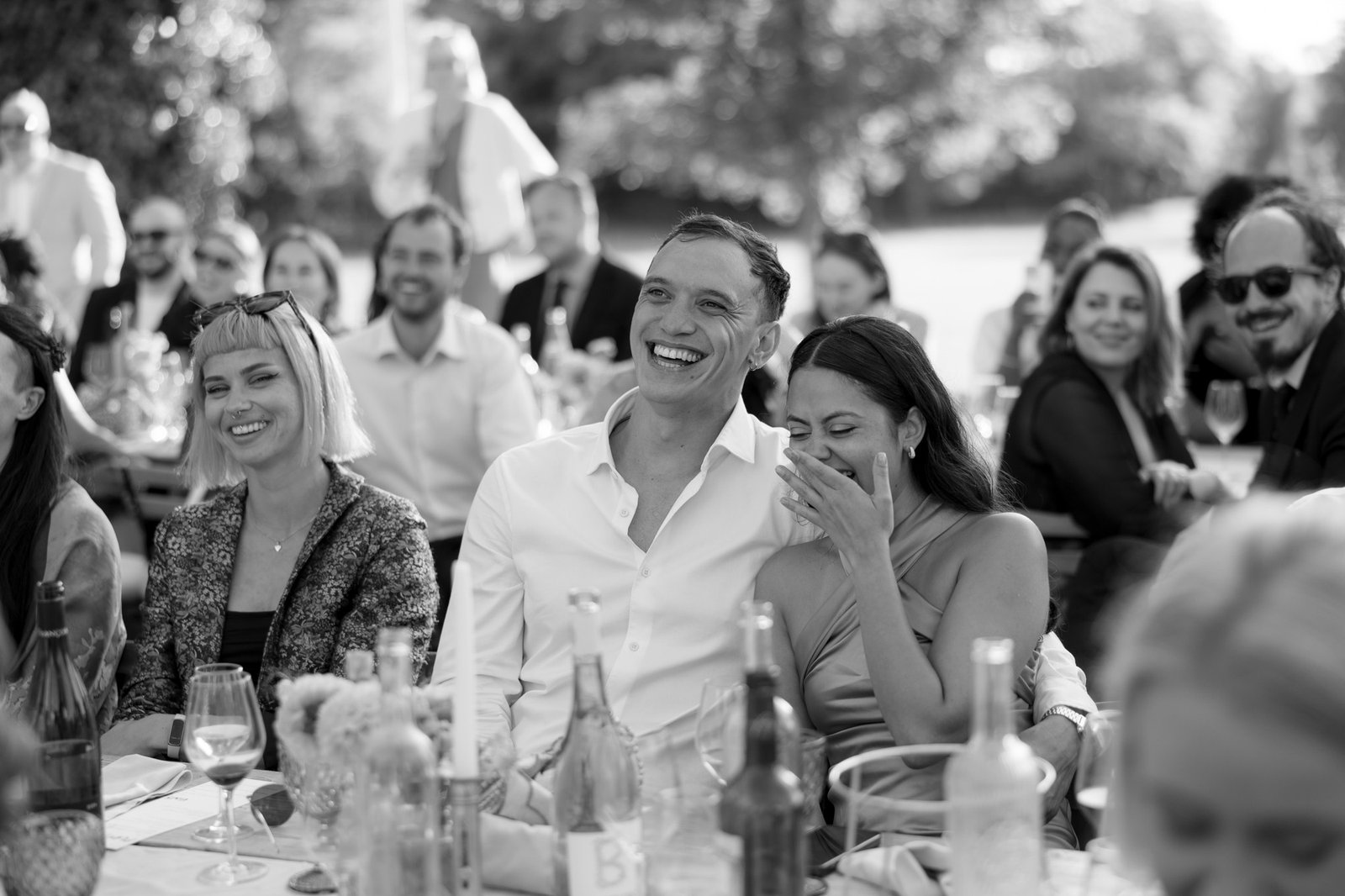 A group of people sit at outdoor tables, smiling and laughing while watching something off-camera at a daytime event.