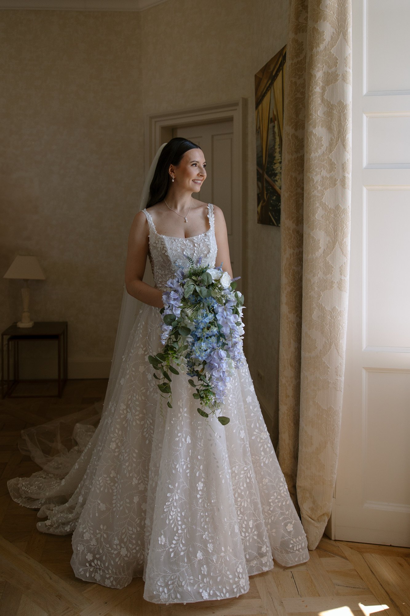 A bride in a white, floral-patterned gown holds a cascading bouquet of blue and white flowers while standing by a window and looking outside. Spring Dordogne Wedding Photographer